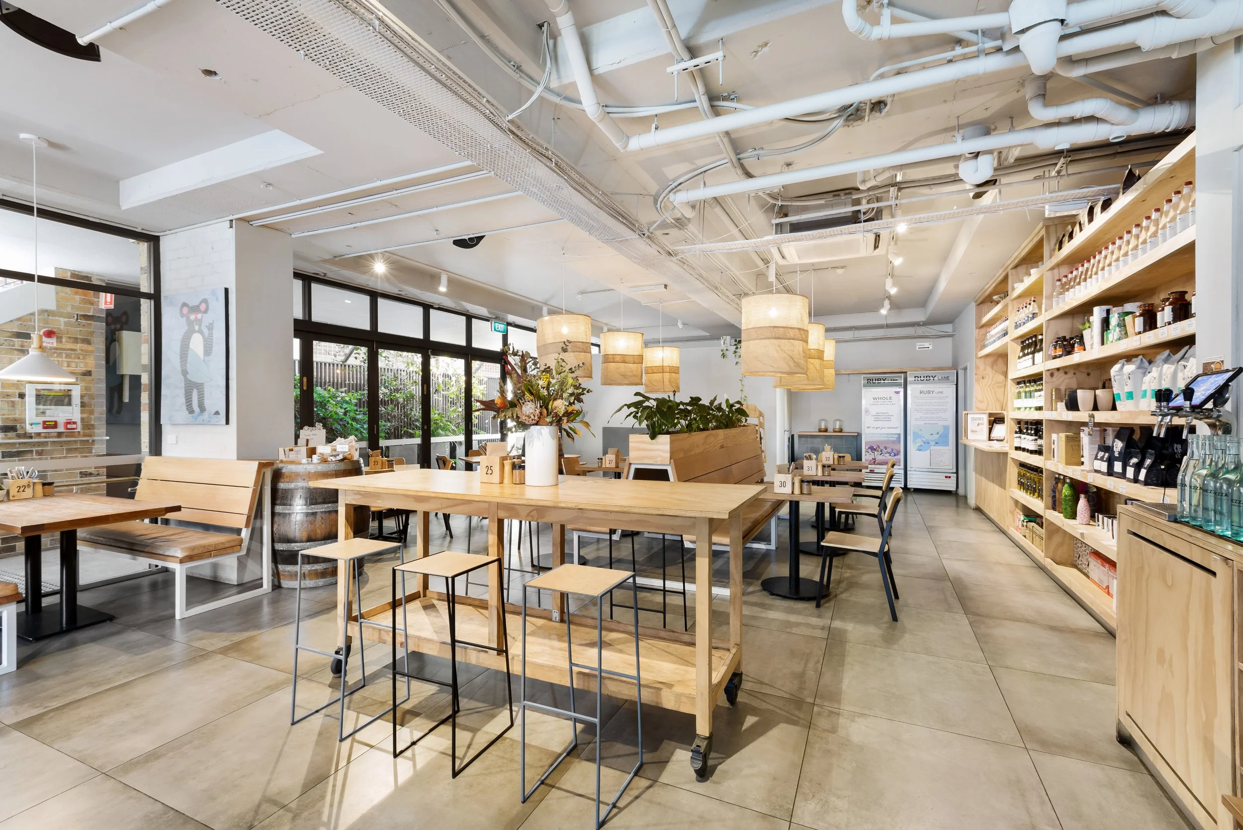 Interior of a modern, well-lit cafe with wooden tables, chairs, and decorative hanging lamps. Green plants and shelves with products are visible, creating a cozy atmosphere.