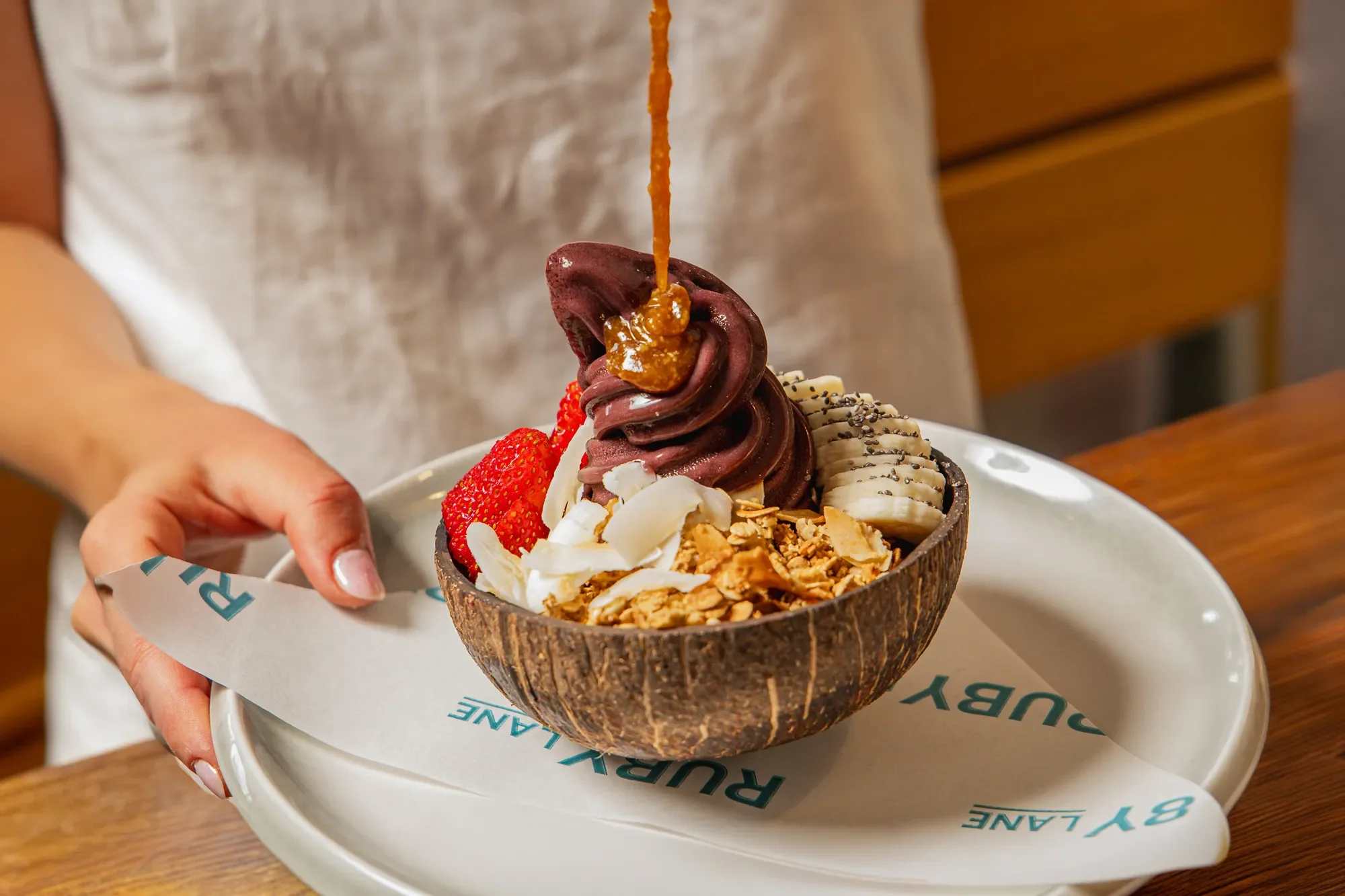 Brown coconut shell bowl filled with frozen yogurt topped with strawberries, banana slices, coconut flakes, granola, and caramel sauce being drizzled on top.