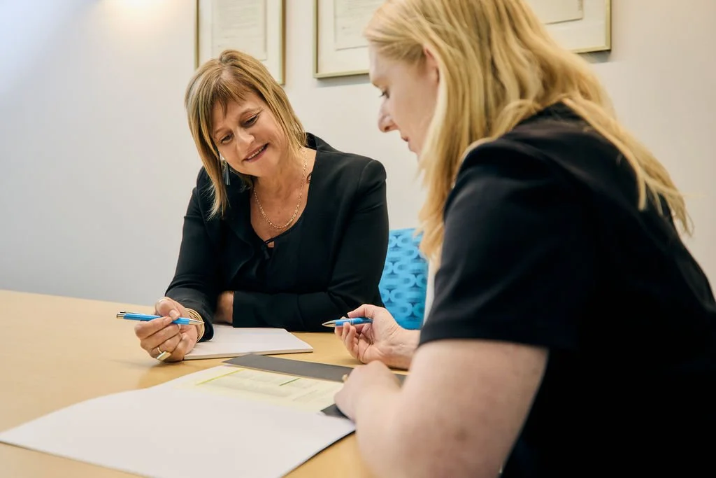 Two women are sitting at a table engaged in a discussion, with papers and pens in front of them, in an office setting.