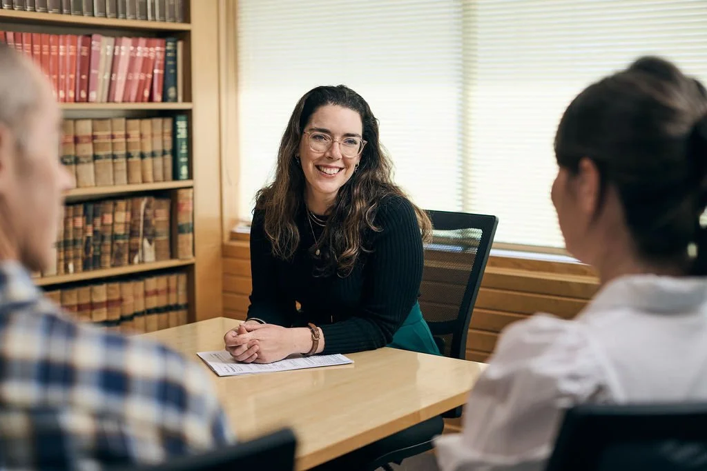 A woman sitting at a table, speaking with two others during a meeting, with bookshelves in the background.