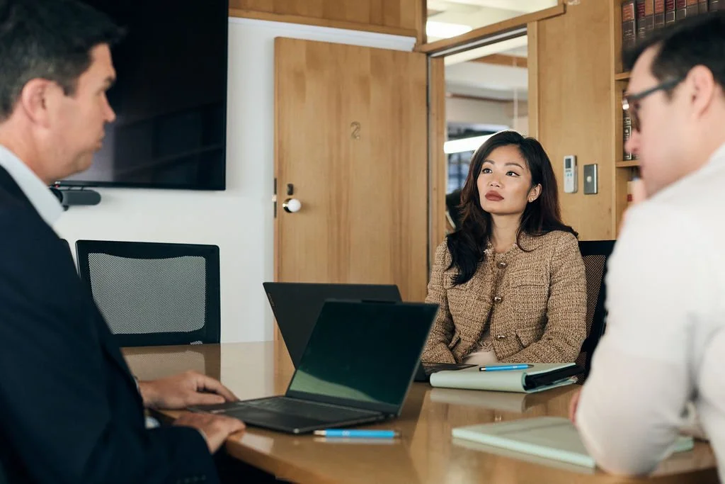 Three people in a business meeting in a conference room with wooden walls, a large TV screen, and a door in the background, with laptops, notebooks, and pens on the table.