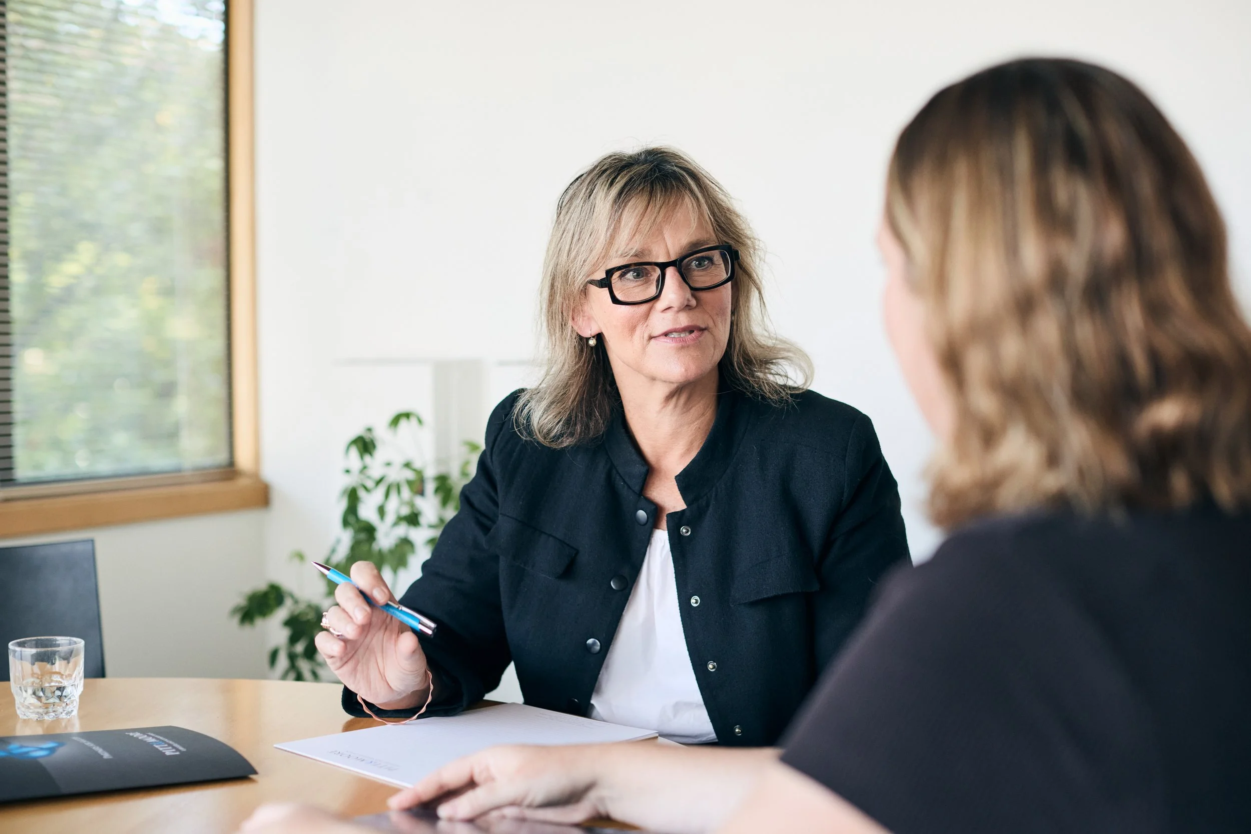 Two women having a conversation at a meeting in an office, one woman with blonde hair and glasses, holding a pen, speaking to another woman with brown hair.