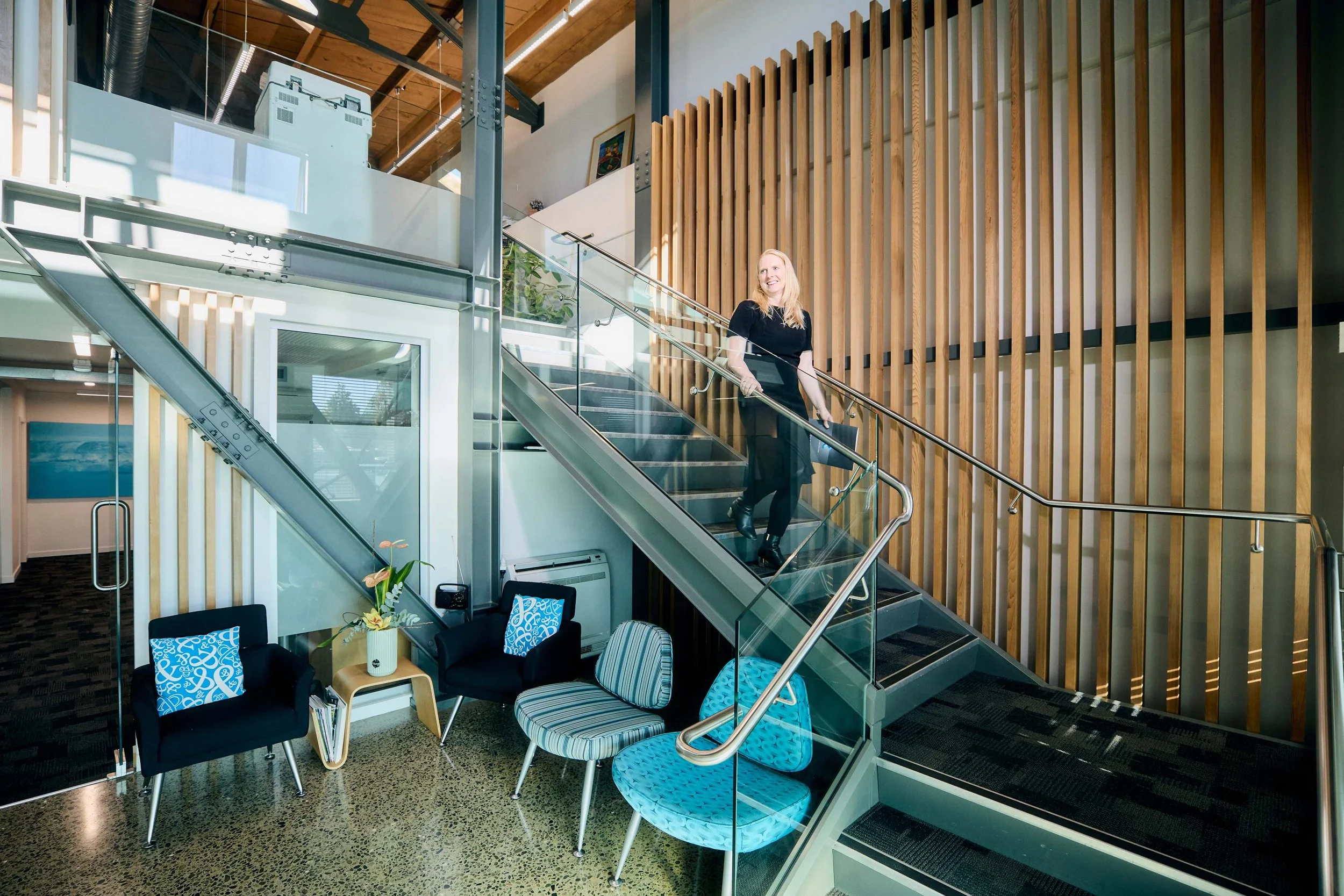 A woman with blonde hair wearing a black outfit, standing on a staircase with glass railing inside a modern building with wood paneling and a high wooden ceiling, smiling while holding a black folder.