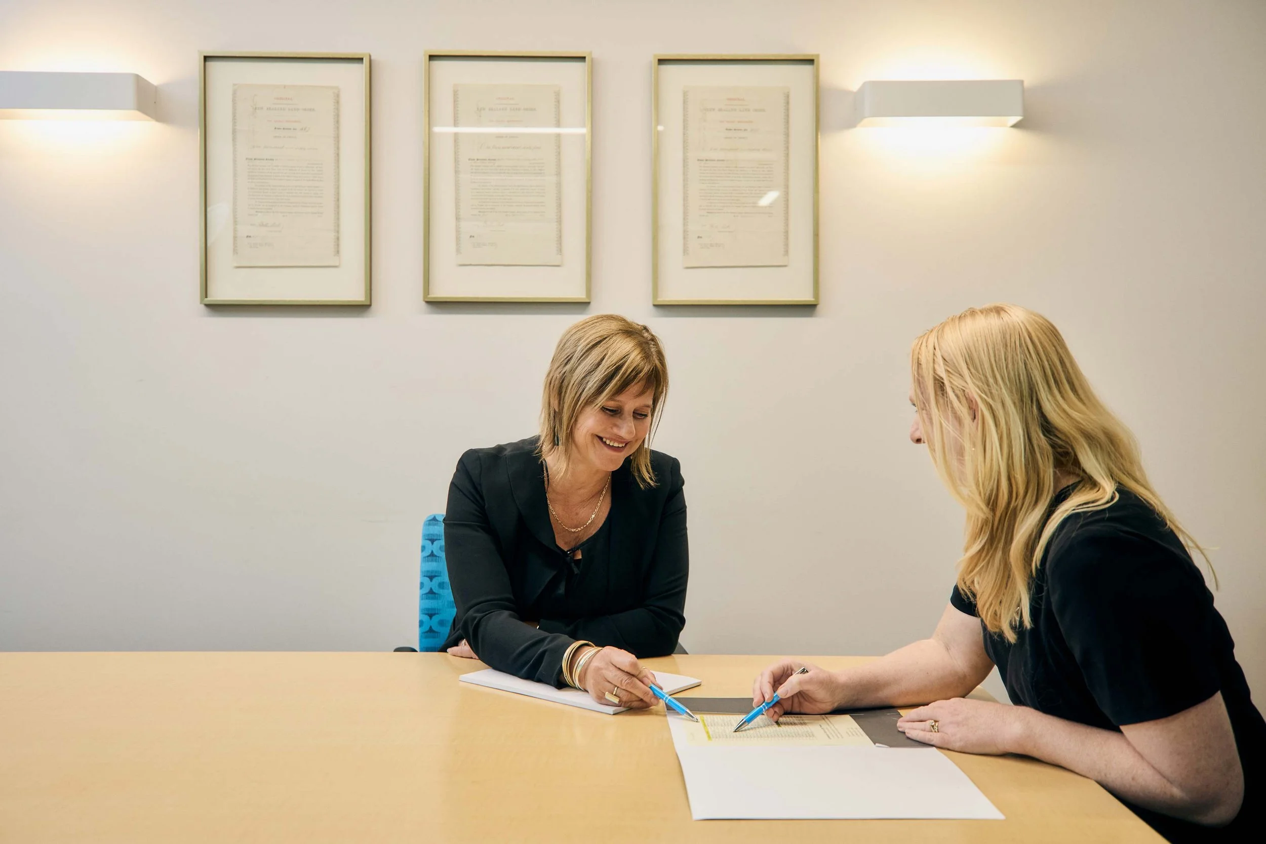 Two women are sitting at a table discussing and reviewing documents in a professional setting. One woman is smiling and holding a pen over a paper, while the other is pointing at the document. The background shows framed certificates or photographs on the wall.