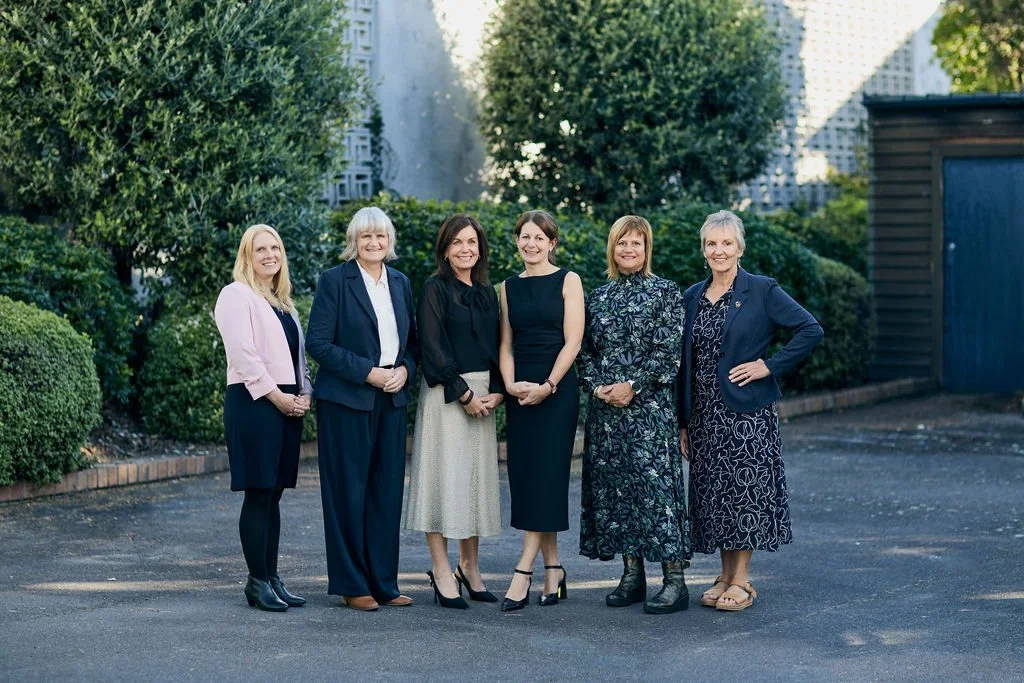 Six women standing outdoors in a row, smiling, with trees and a building in the background, dressed in professional attire.