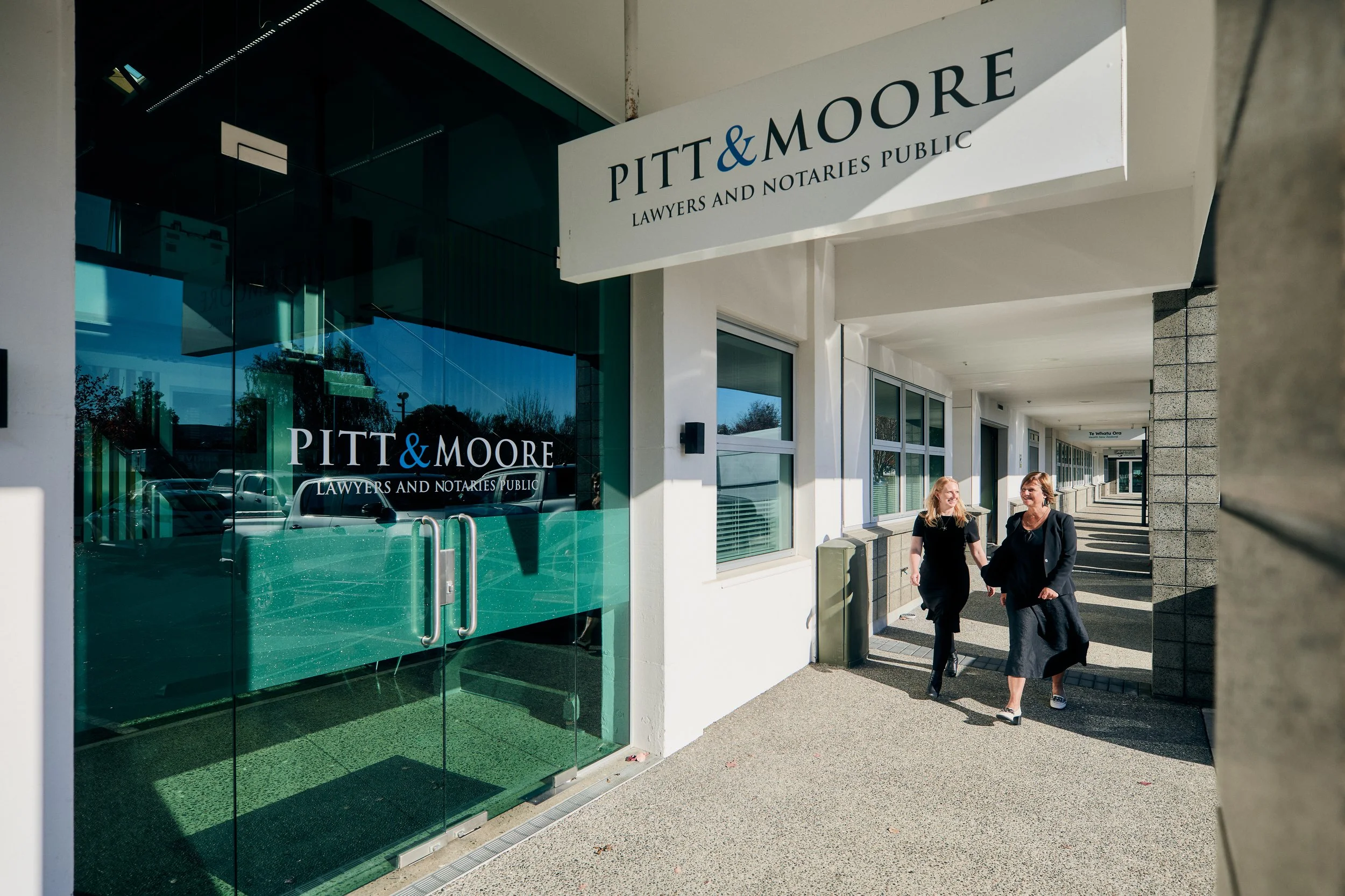 Two women walking and talking along a sidewalk outside a law office with a sign reading 'Pitt & Moore Lawyers and Notaries Public' and large glass windows reflecting nearby buildings and cars.