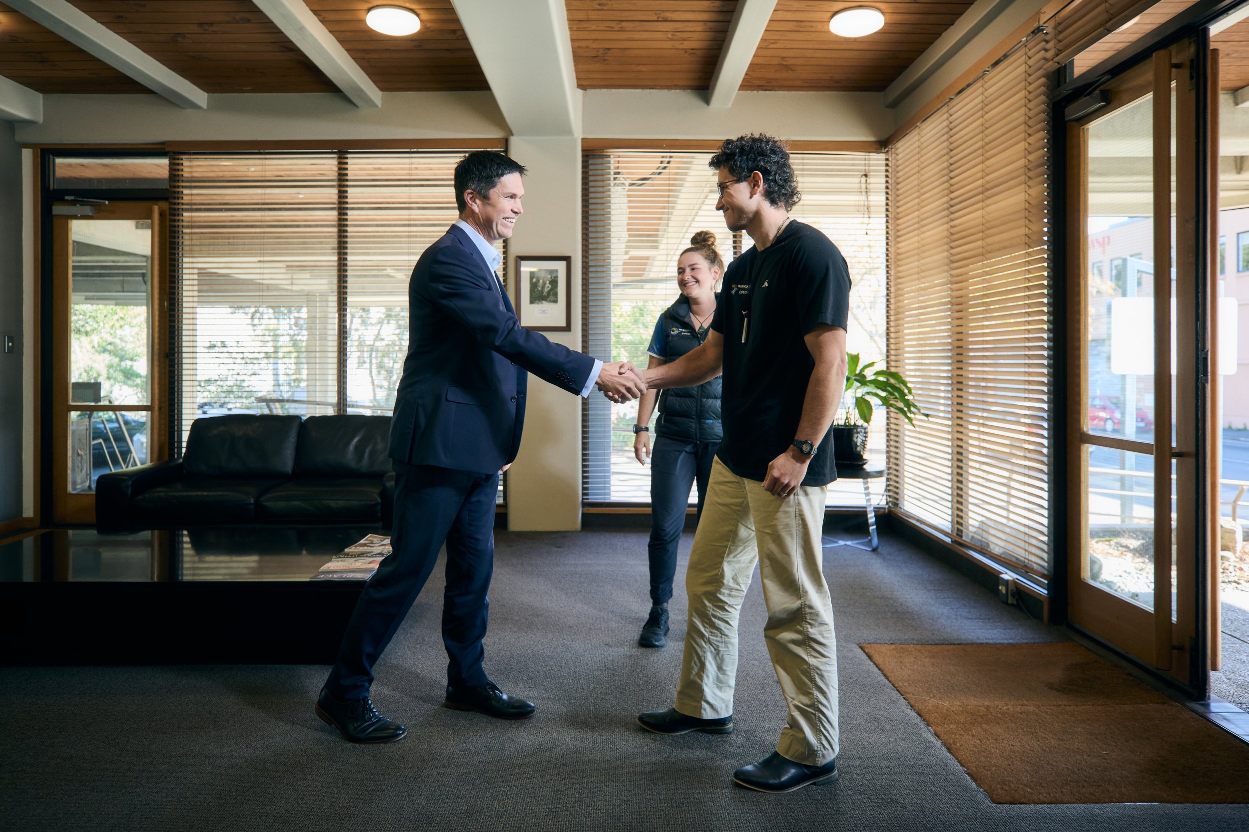 Man in business attire shaking hands with a young man in casual clothes while a woman stands smiling in the background inside an office with wooden ceilings and blinds covering large windows.