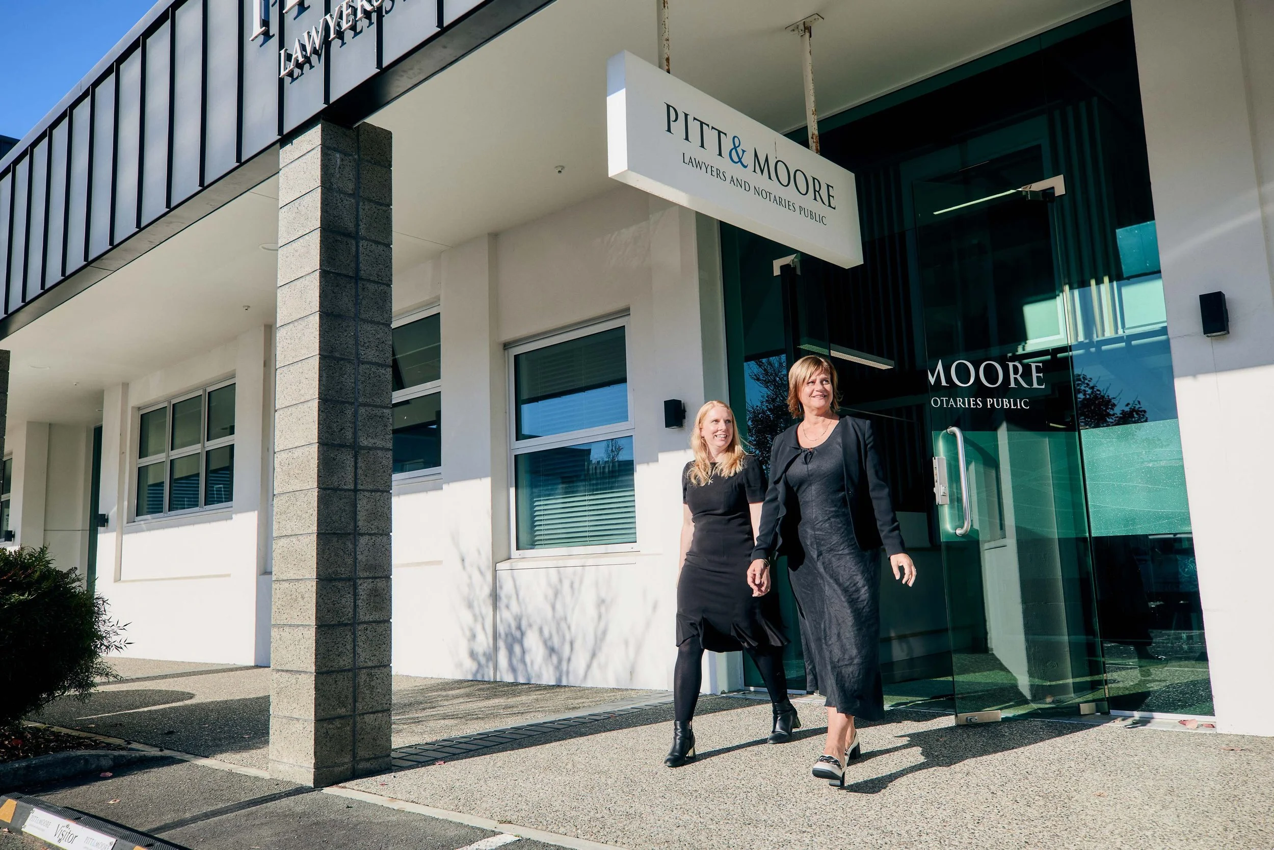 Two women walking out of a building with a sign that reads "Pitt & Moore Lawyers and Notaries Public." The building has a modern exterior with large glass doors and windows, and the women are dressed in black outfits.