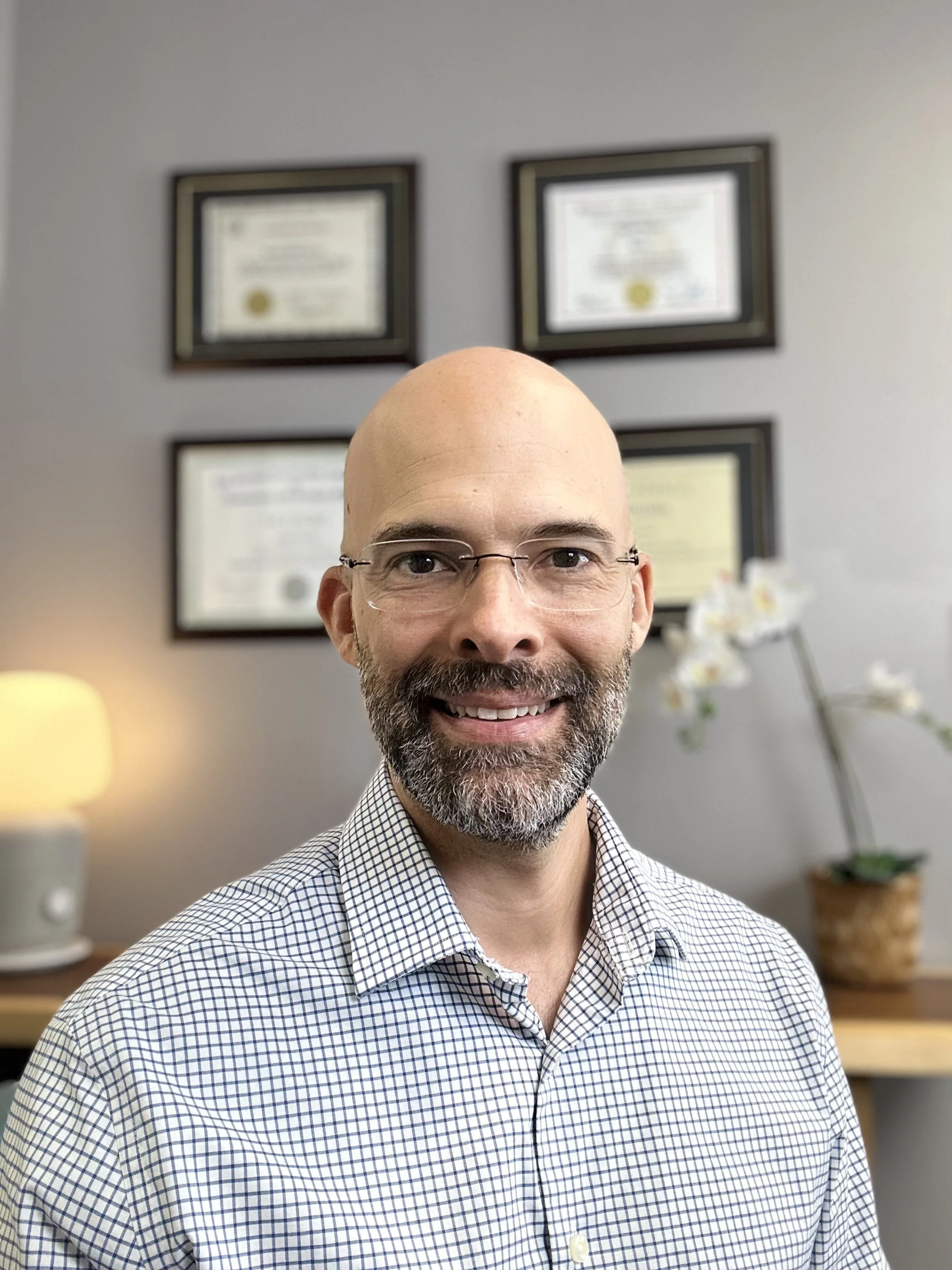 Dr. Justin Garcia smiling in a professional setting with framed diplomas on the wall and an orchid plant in the background.