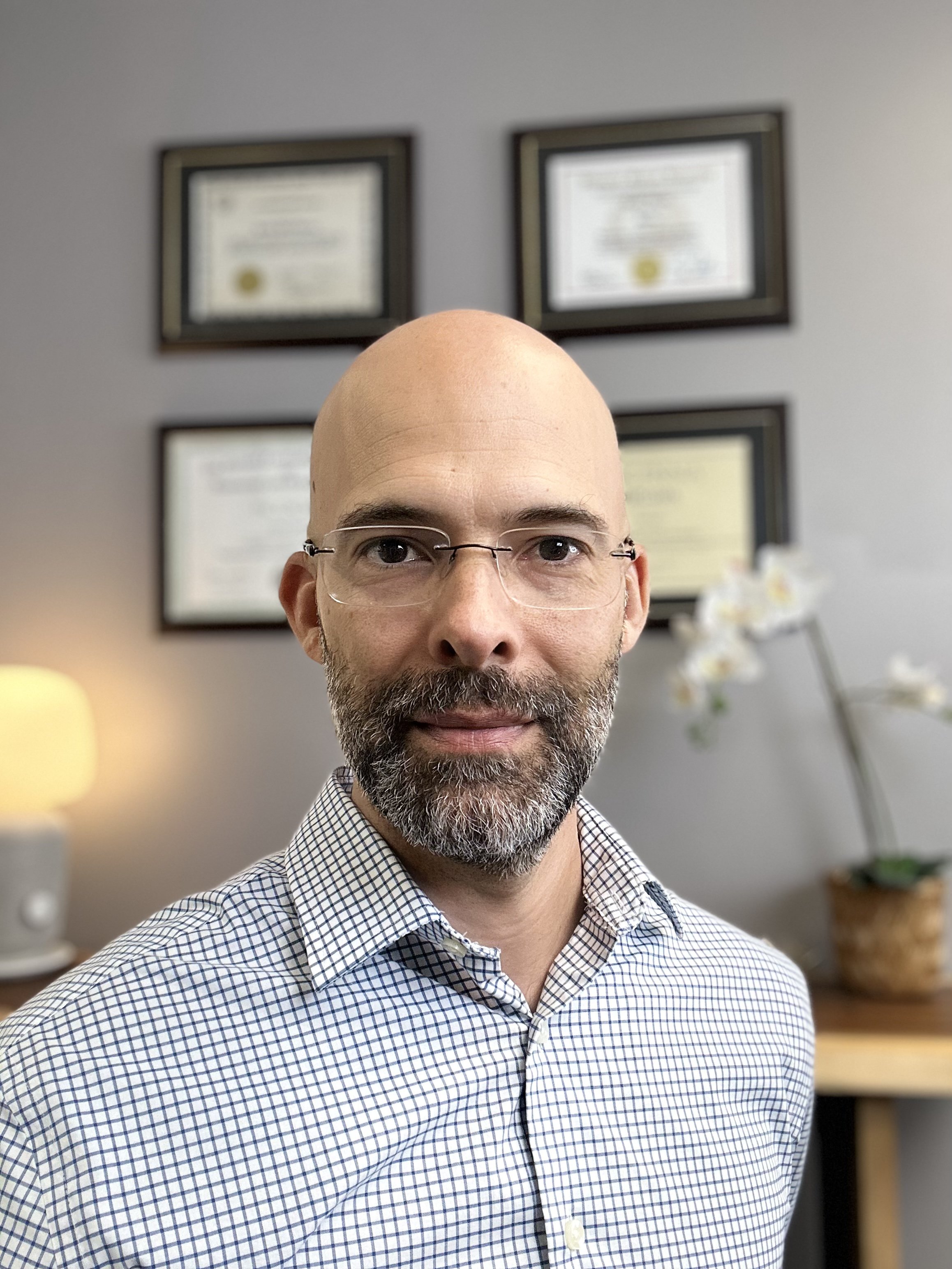 Dr. Justin Garcia smiling in a professional setting with framed diplomas on the wall and an orchid plant in the background.
