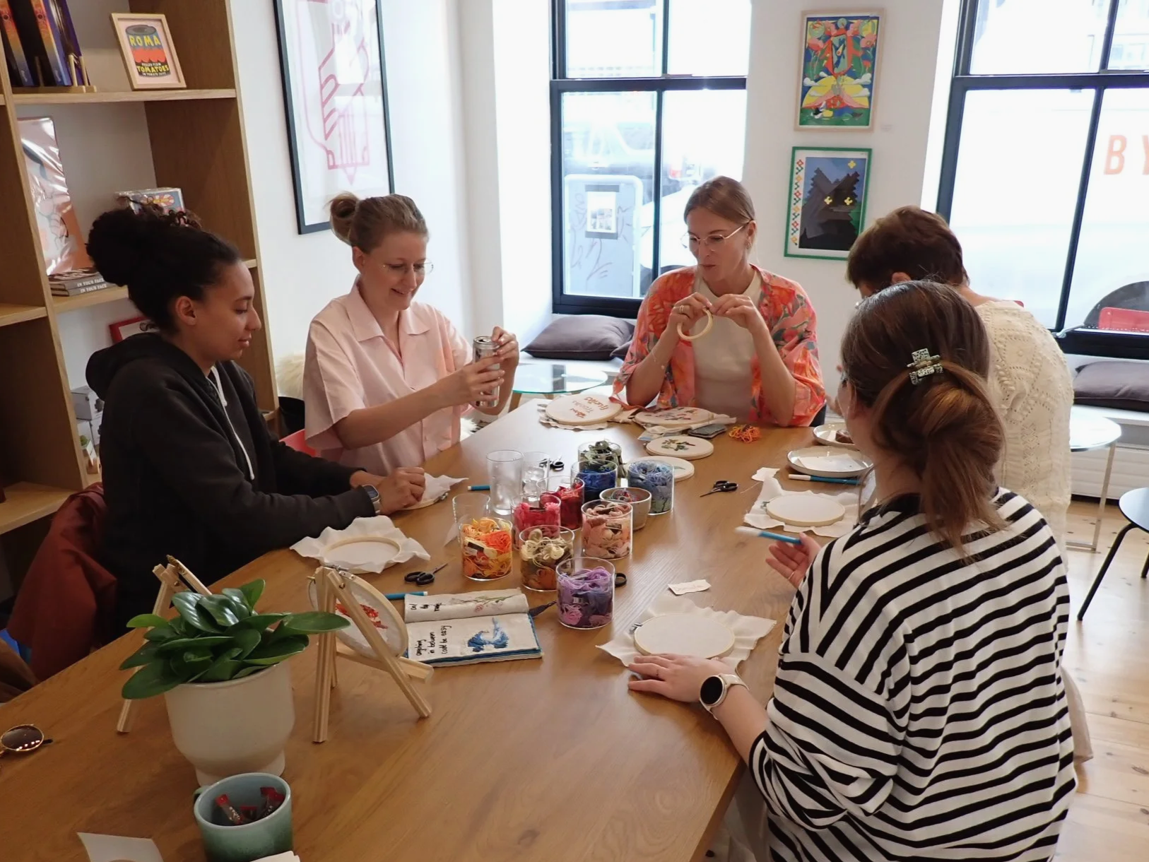 A group of six women sitting around a table engaged in embroidery or craft activities in a well-lit room with large windows and colorful art on the walls. The table is filled with thread spools, scissors, and embroidery hoops, and there is a potted p