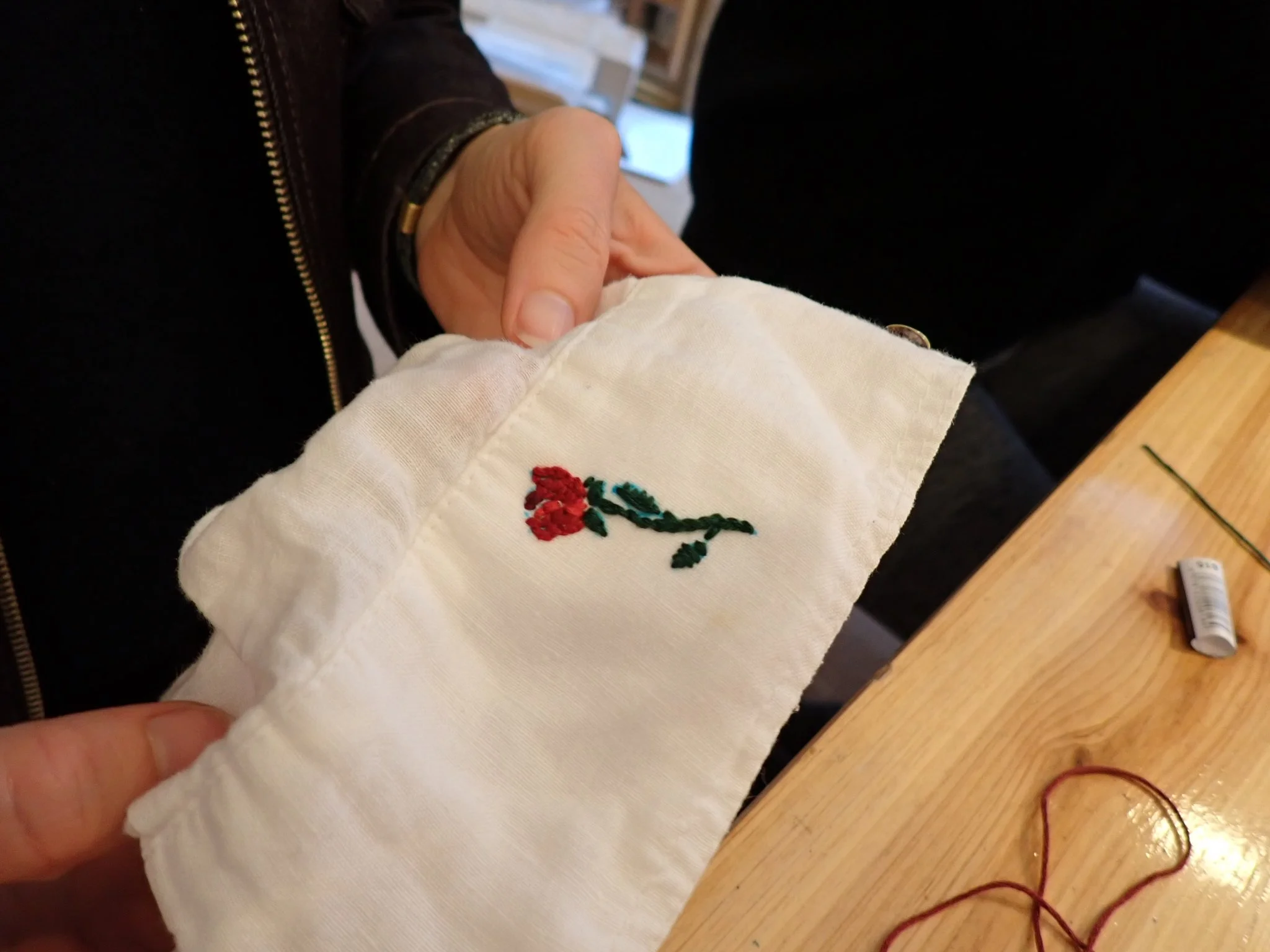 Close-up of a person's hand holding a white cloth with embroidery of a red flower and green leaves, with another person beside them wearing a black shirt and a brown jacket with a zipper. The background shows a wooden table, a small tube of glue, and