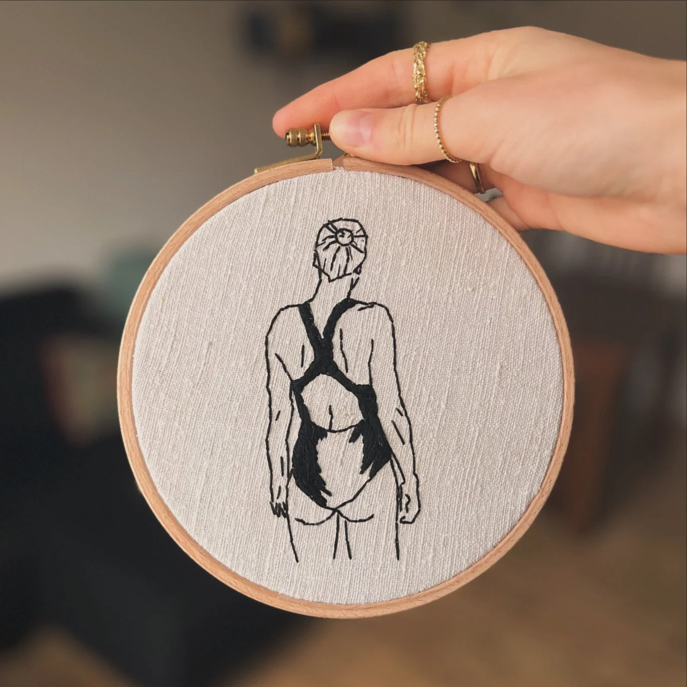 A hand holding an embroidery hoop with a drawing of a woman in a swimsuit, viewed from behind.