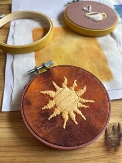 Embroidery hoop with a yellow sun design on orange fabric, jewelry box with a white flower-shaped brooch, and a container with soaps or skincare products.