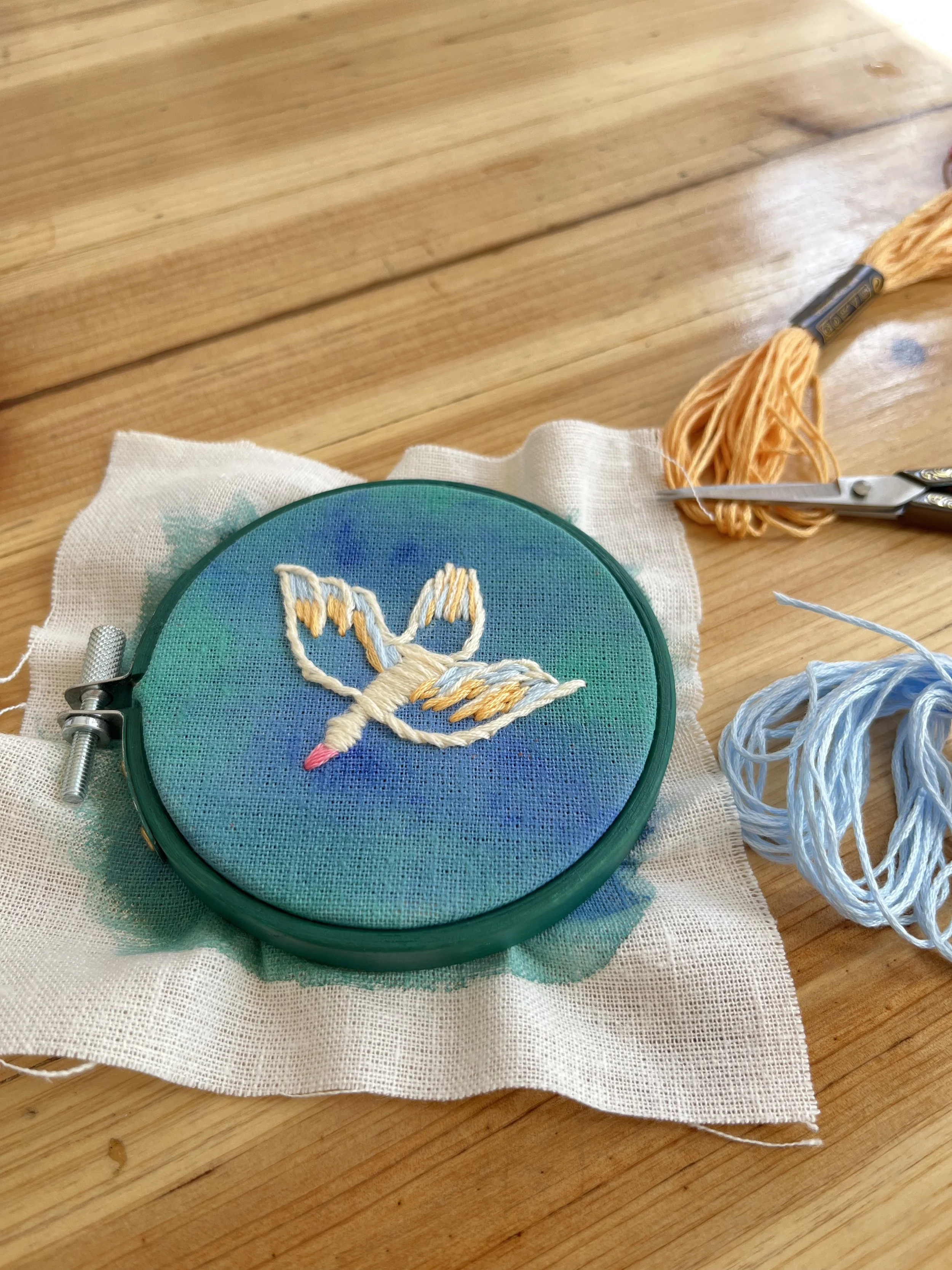 Embroidery hoop with a bunny design in progress, set on a piece of fabric on a wooden table, with scissors, thread, and floss nearby.