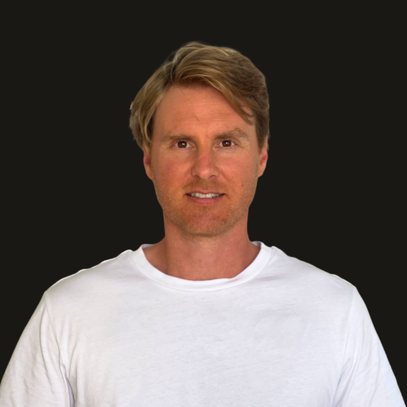 A man with light brown hair and a slight beard, wearing a white t-shirt, smiling against a black background.