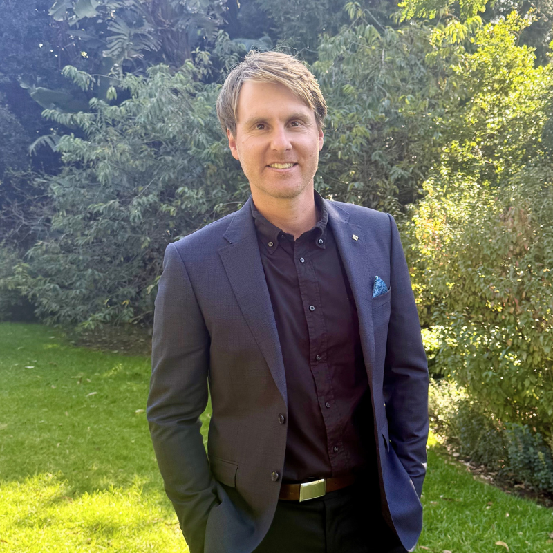 A man in a navy blazer and black shirt standing outdoors on grass with greenery in the background.