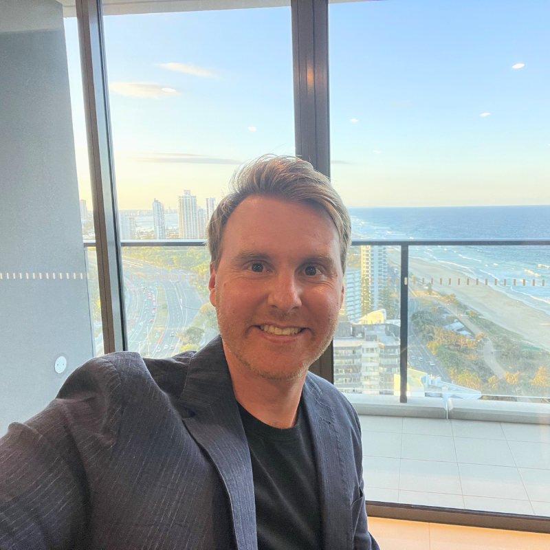 A man smiling for a selfie in front of a large window overlooking a cityscape, ocean, and beach with tall buildings, a highway, greenery, and a clear sky in the background.