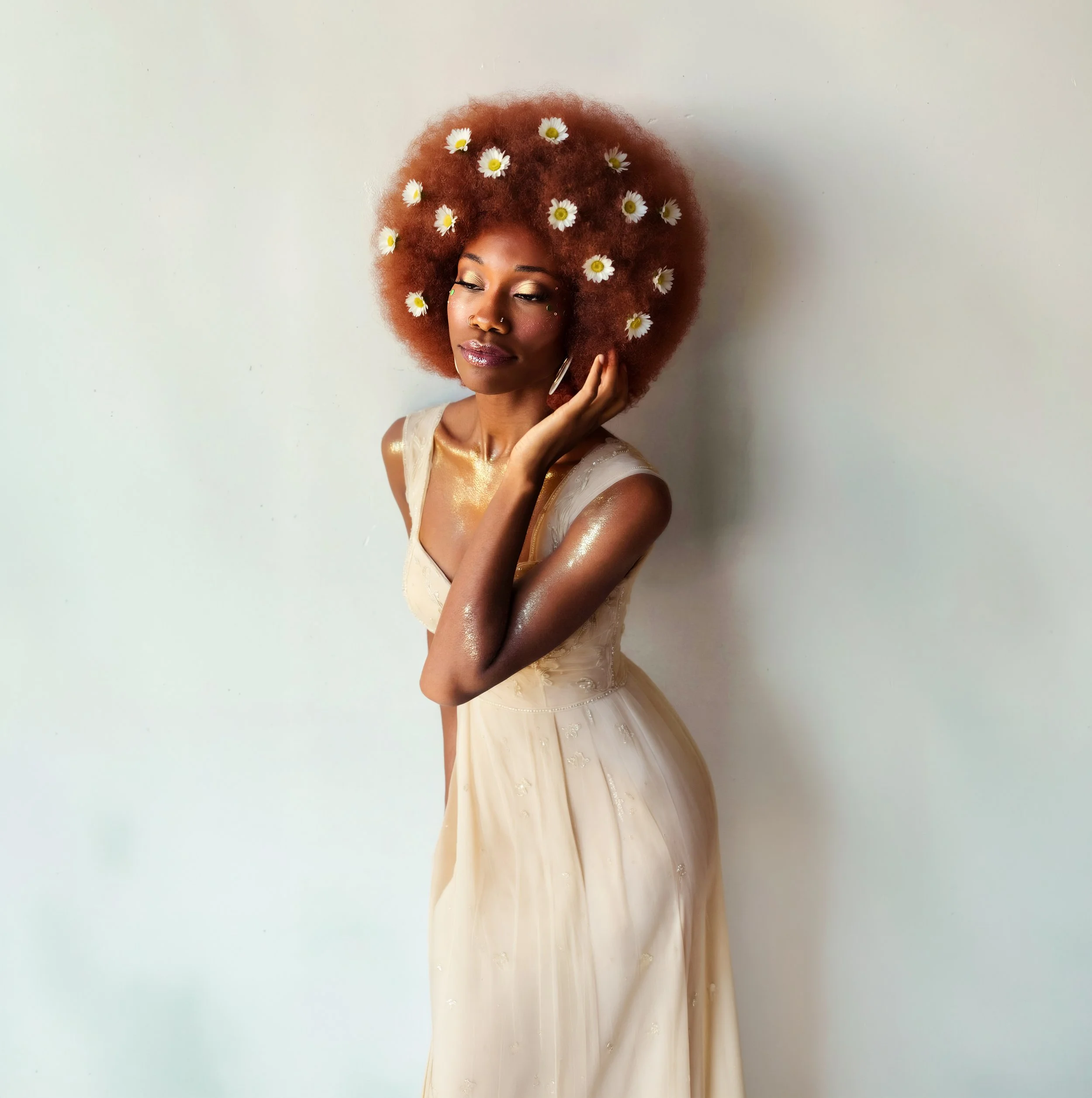 Young woman with curly red hair adorned with daisies, wearing a cream-colored dress, standing against a plain white wall.