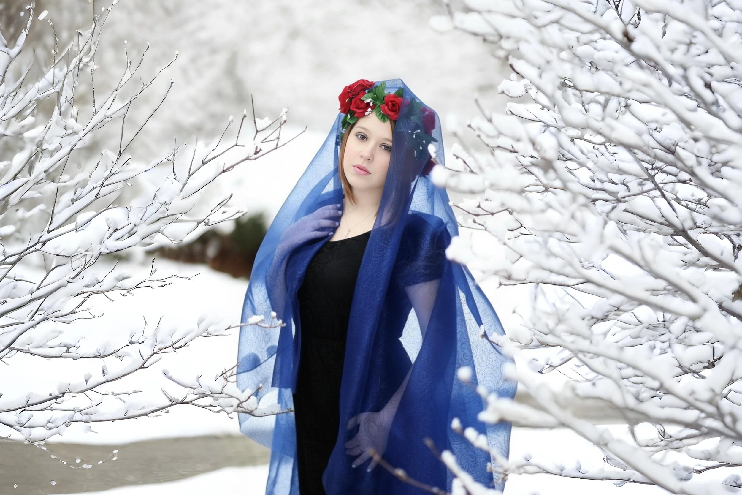 A woman in a black dress with a blue sheer shawl and a red rose headpiece stands in a snowy winter landscape surrounded by snow-covered branches.