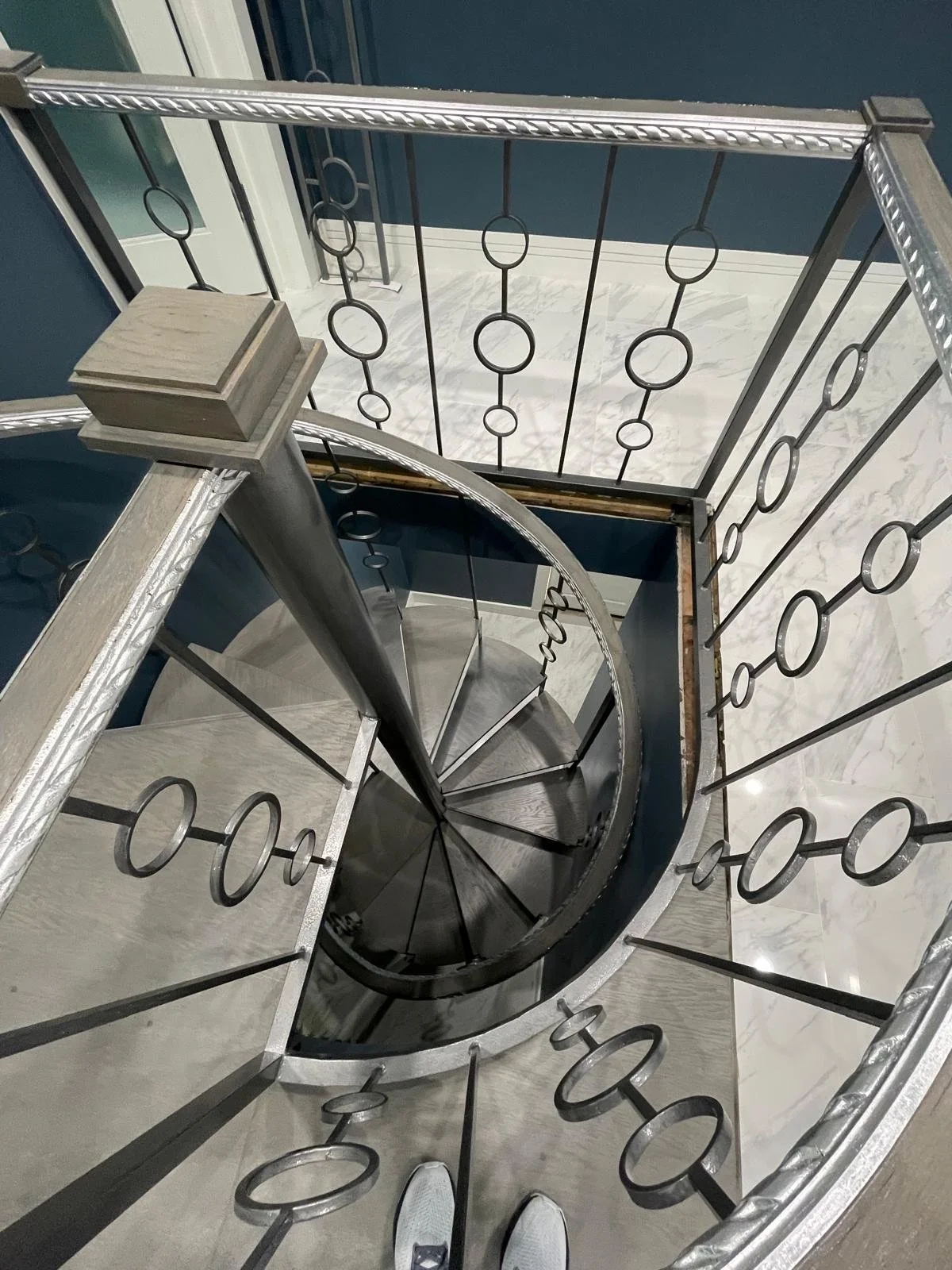 View from the top of a spiral staircase with metal railings featuring circular designs, leading down to a marble-floored area. The staircase is enclosed with blue walls and some white moldings.