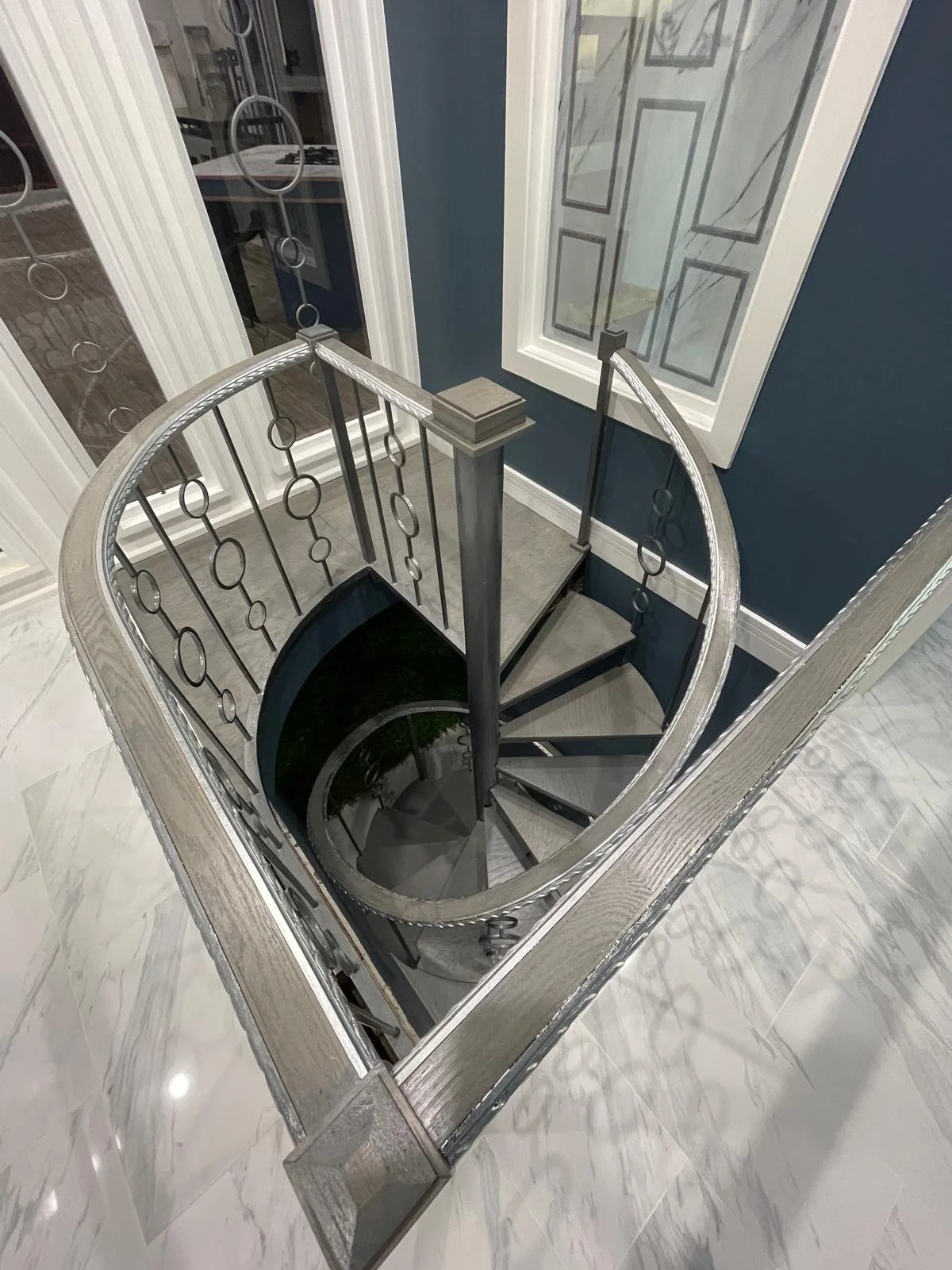 Spiral staircase with metal railings seen from above in a hallway, next to a window. The staircase has dark steps and a decorative railing design.