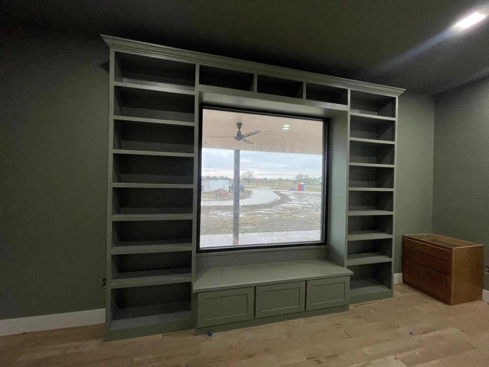 Empty built-in bookshelf with gray finish surrounding a window in a room with hardwood flooring.