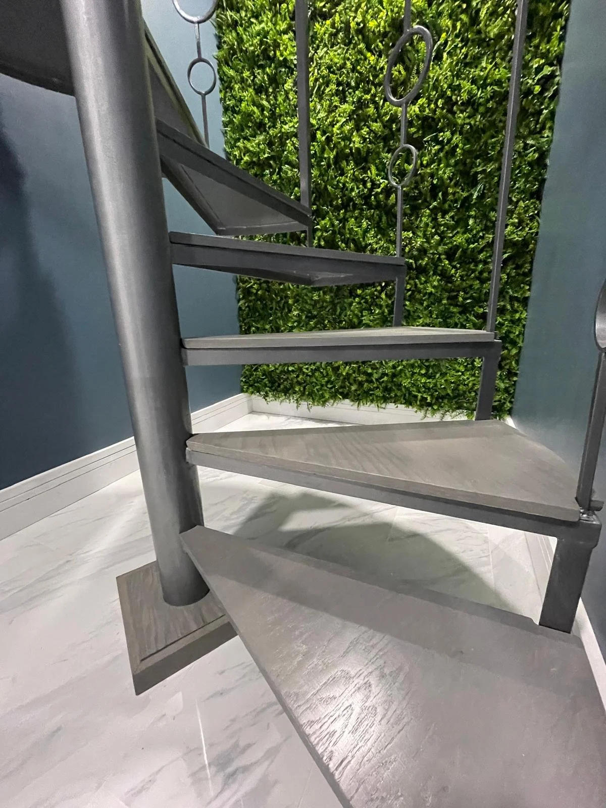 Close-up of a modern metal spiral staircase with textured steps, against a background of a green leafy wall and blue walls.