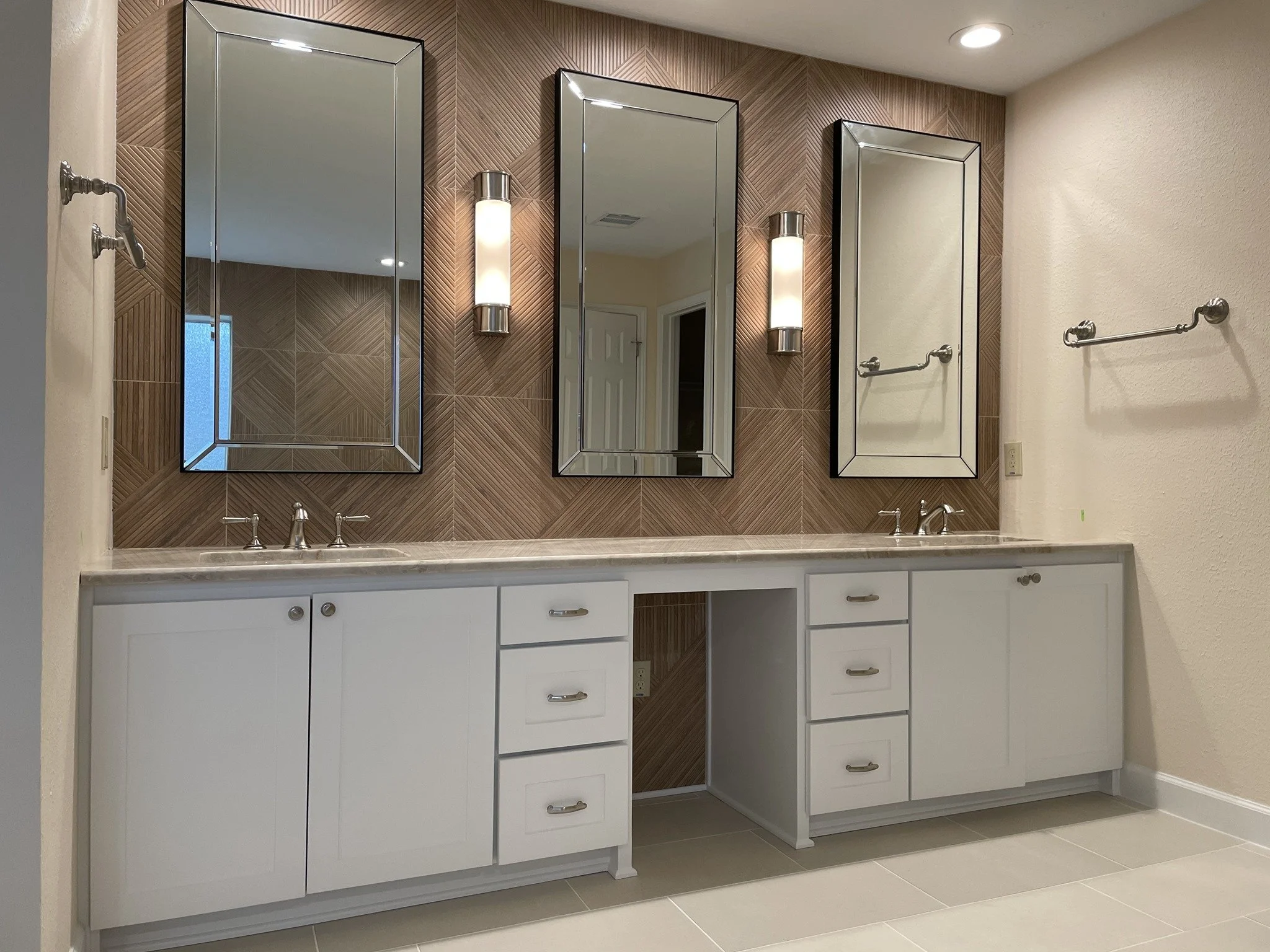 A bathroom double vanity with three large rectangular mirrors, wall sconces, white cabinets, a marble countertop, and a wood-tiled accent wall.