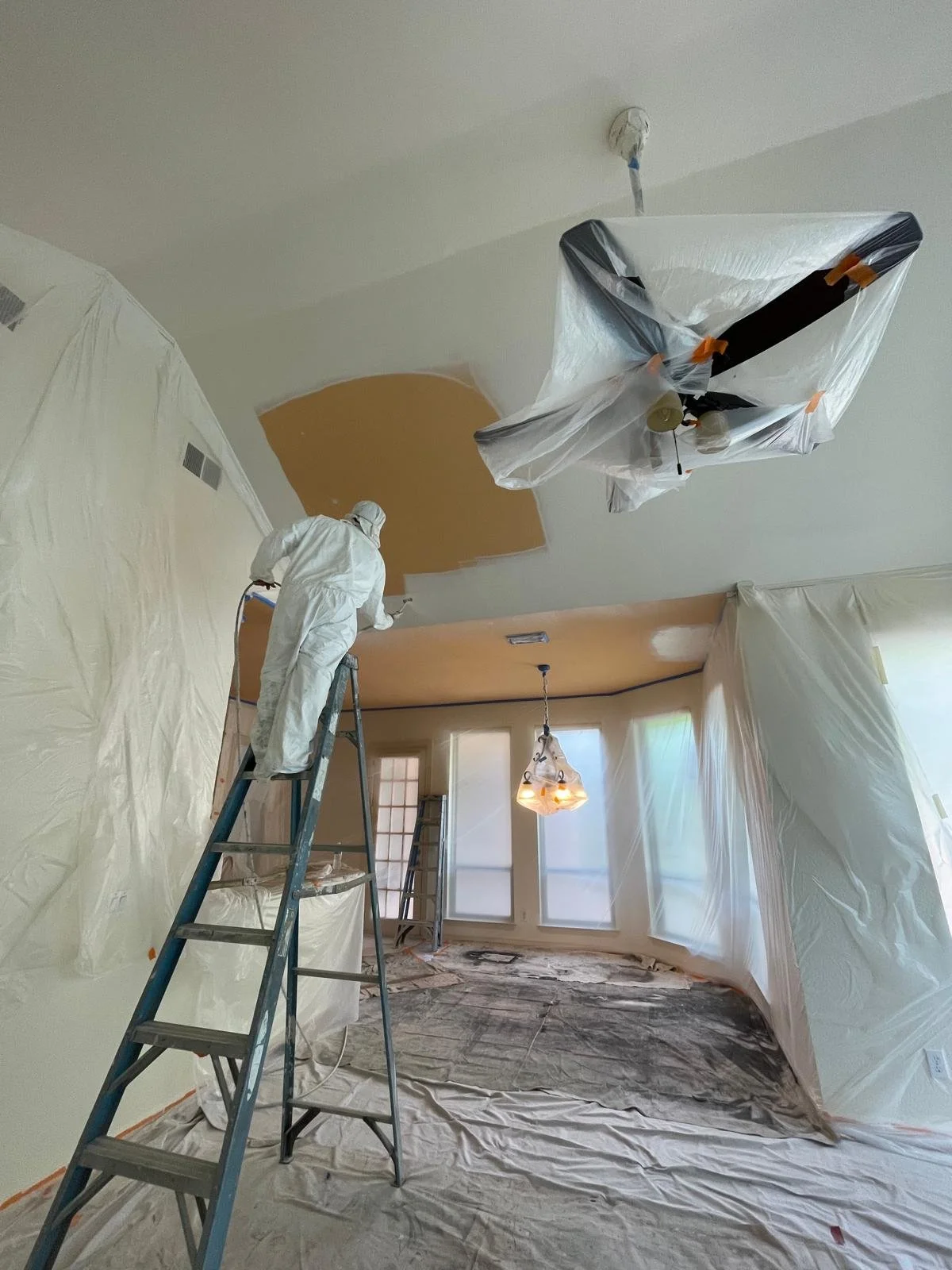 Person in white protective suit painting a ceiling, with walls covered in plastic sheeting to protect from paint splatters, and ladders in a room with large windows.