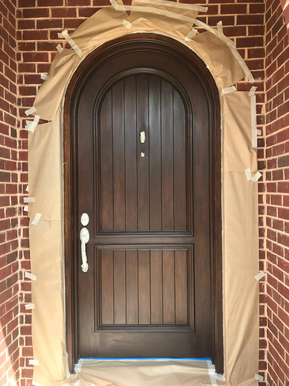 A dark brown arched wooden door surrounded by brick wall, with masking paper and tape around the edges, indicating it is under renovation or painting.