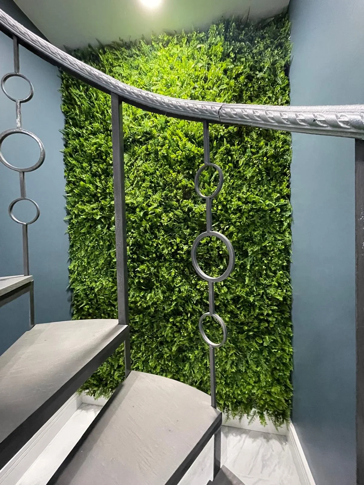Interior staircase with metal handrail featuring circular designs, leading past a green artificial plant wall on the back wall, with gray walls on either side.