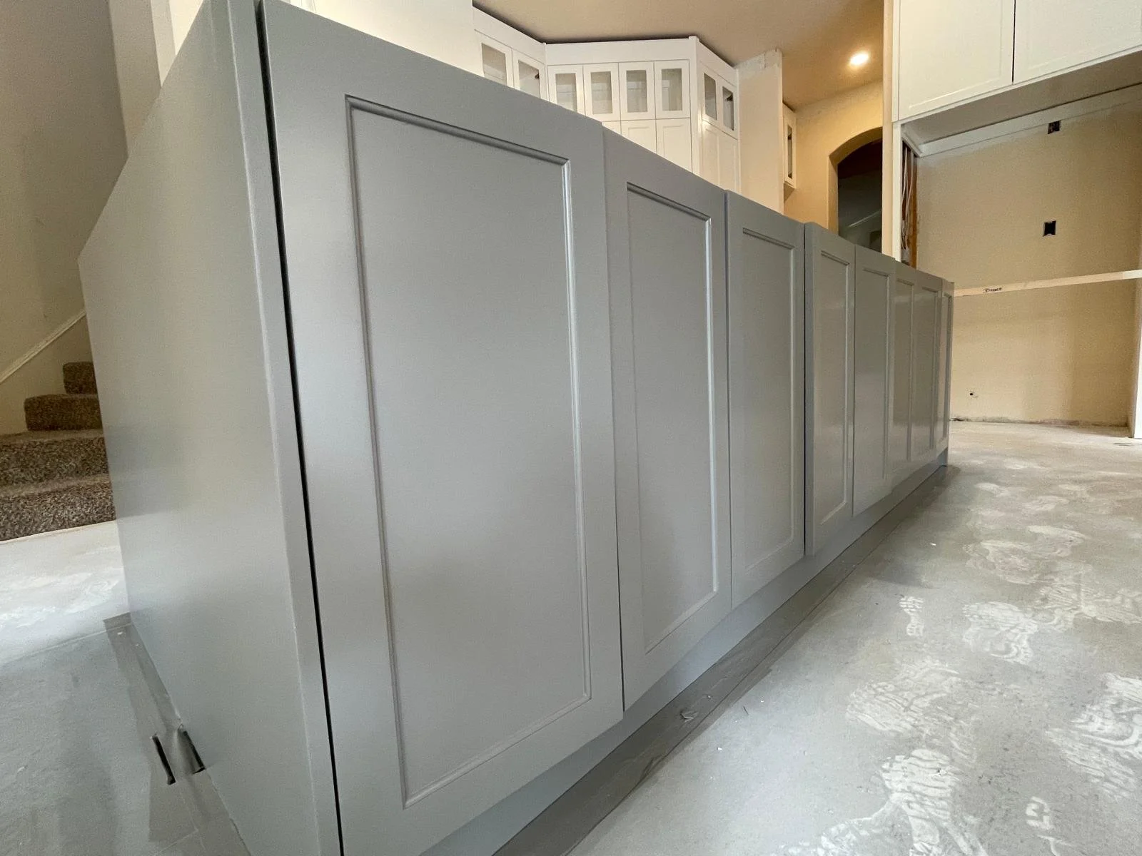A long gray kitchen island with panel details, under construction in a bright room with white cabinetry and unfinished walls.