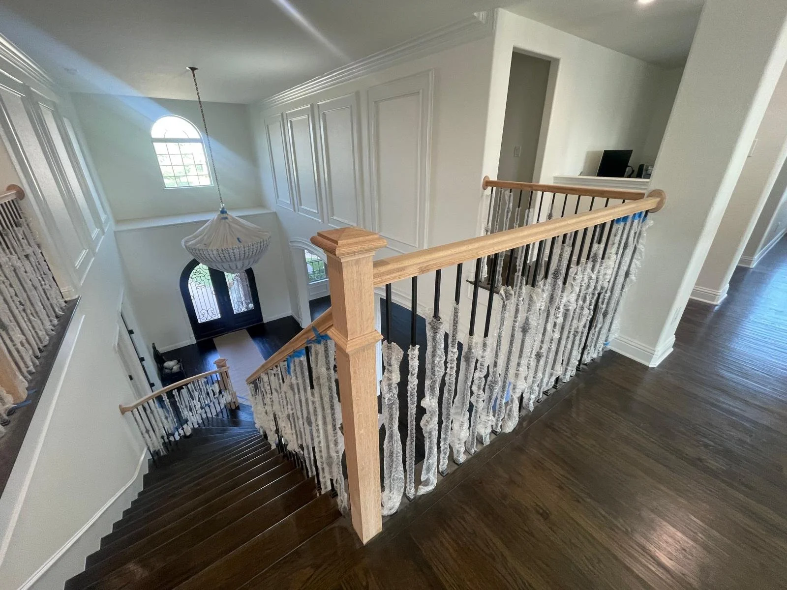 View of an indoor staircase with dark wooden steps and white and black iron balusters. A wooden handrail runs along the staircase and the hallway. The space has white walls, crown molding, and a large window with a semi-circular top near the entrance