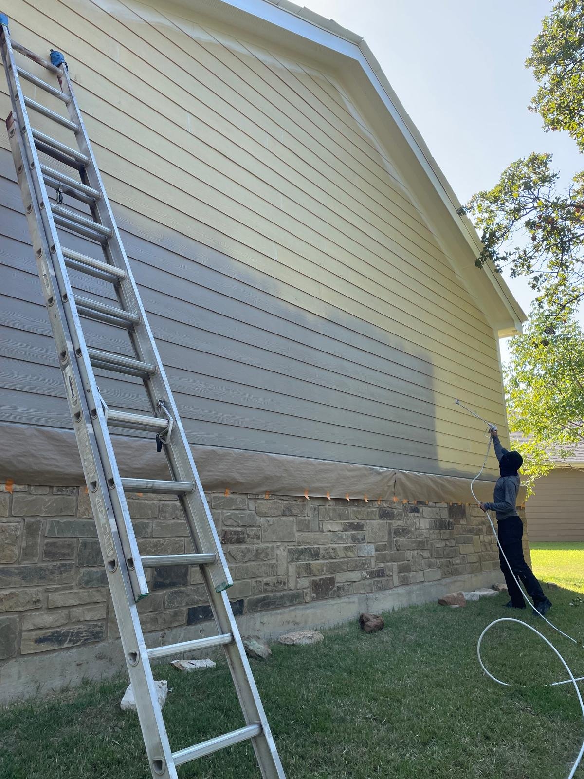 Person power washing the side of a house with beige siding, a ladder leaning against the house, and stones around the foundation.