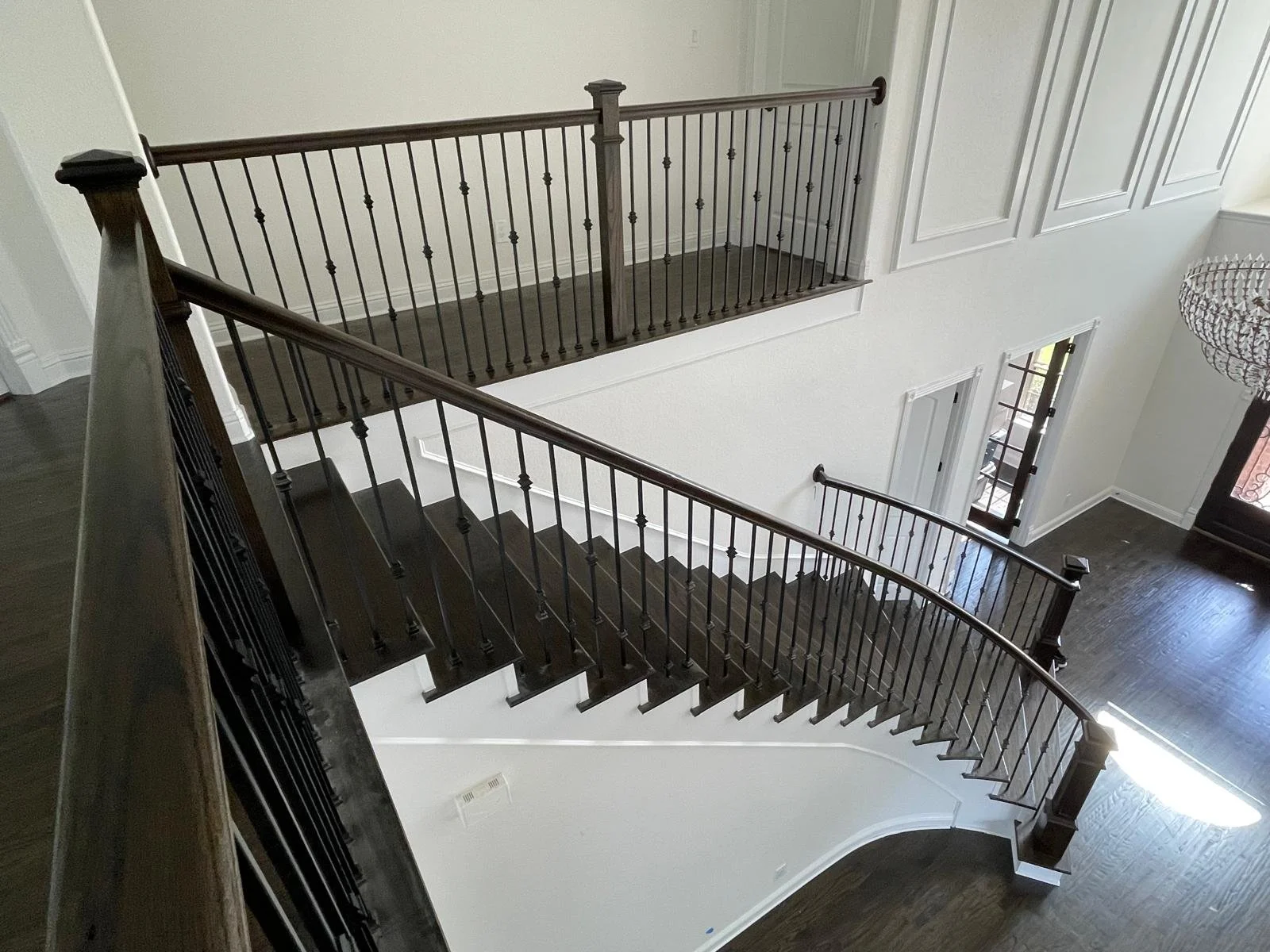 Interior view of a staircase with black wooden steps and black metal railings, leading from the second floor to the first floor, with a door and windows visible at the bottom of the stairs.
