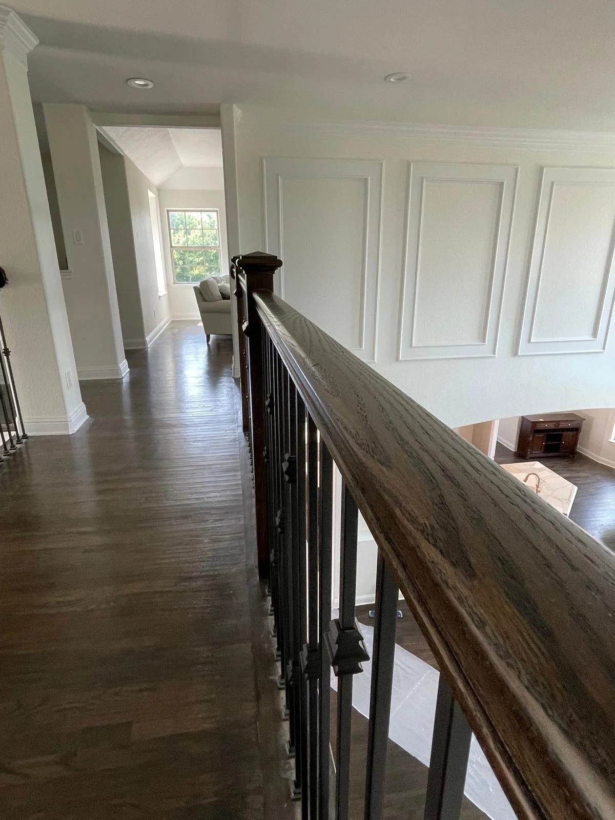 Interior view of a second-floor hallway with dark hardwood flooring, white walls with decorative molding, and a wooden handrail on iron balusters. At the end of the hall, there is a seating area with a chair near a window, with a glimpse of another r