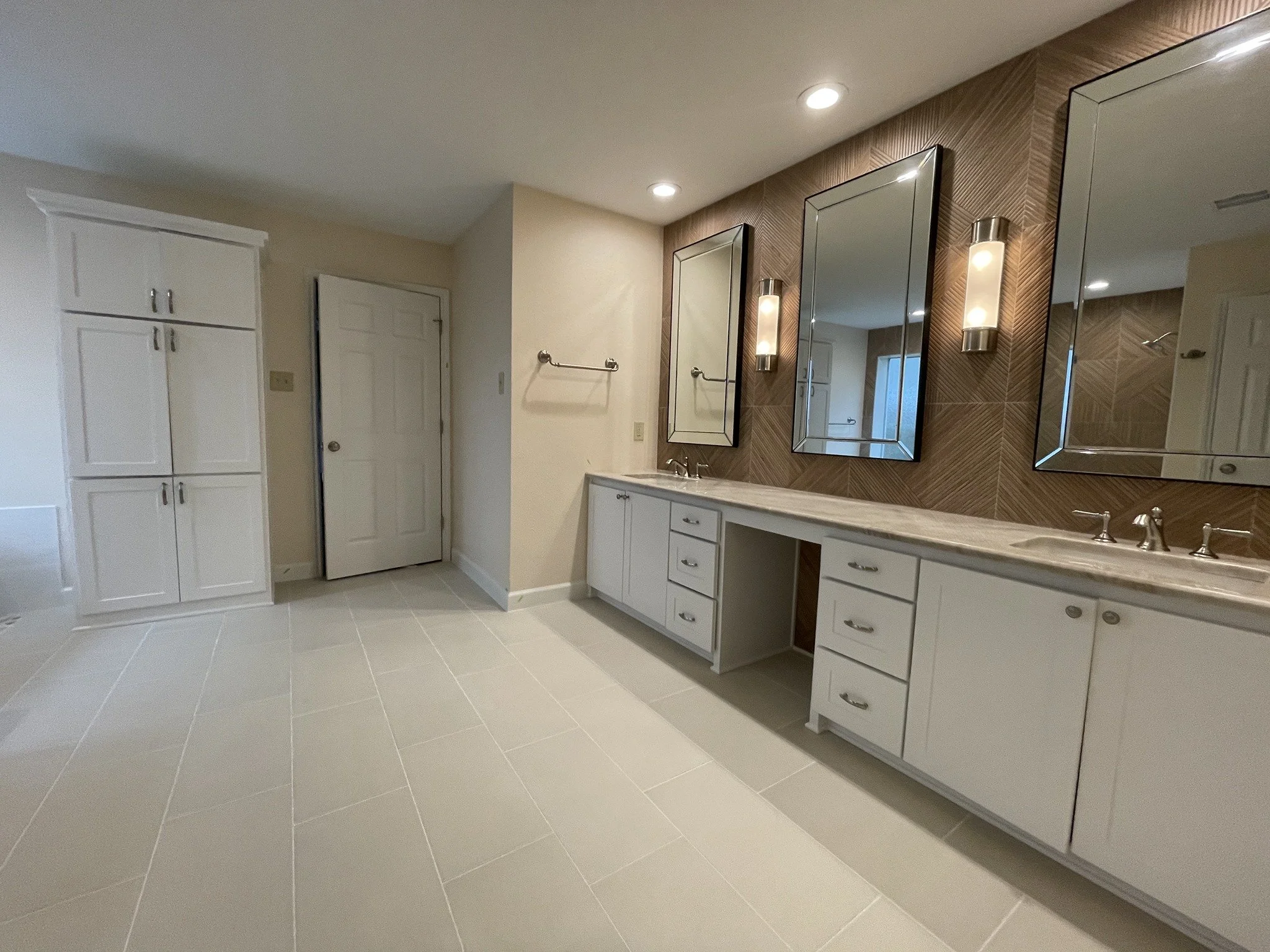 Bathroom with white cabinets, three mirrors, two wall lights, beige tile floor, and a brown pattern wall behind the mirrors.