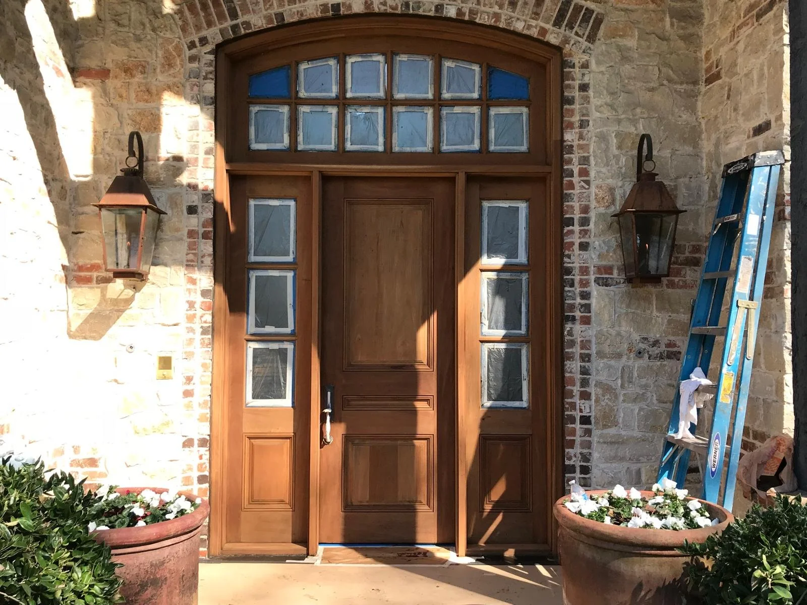 Wooden front door with glass panels on a brick wall, flanked by two lantern-style lights, with potted plants and a blue ladder nearby.