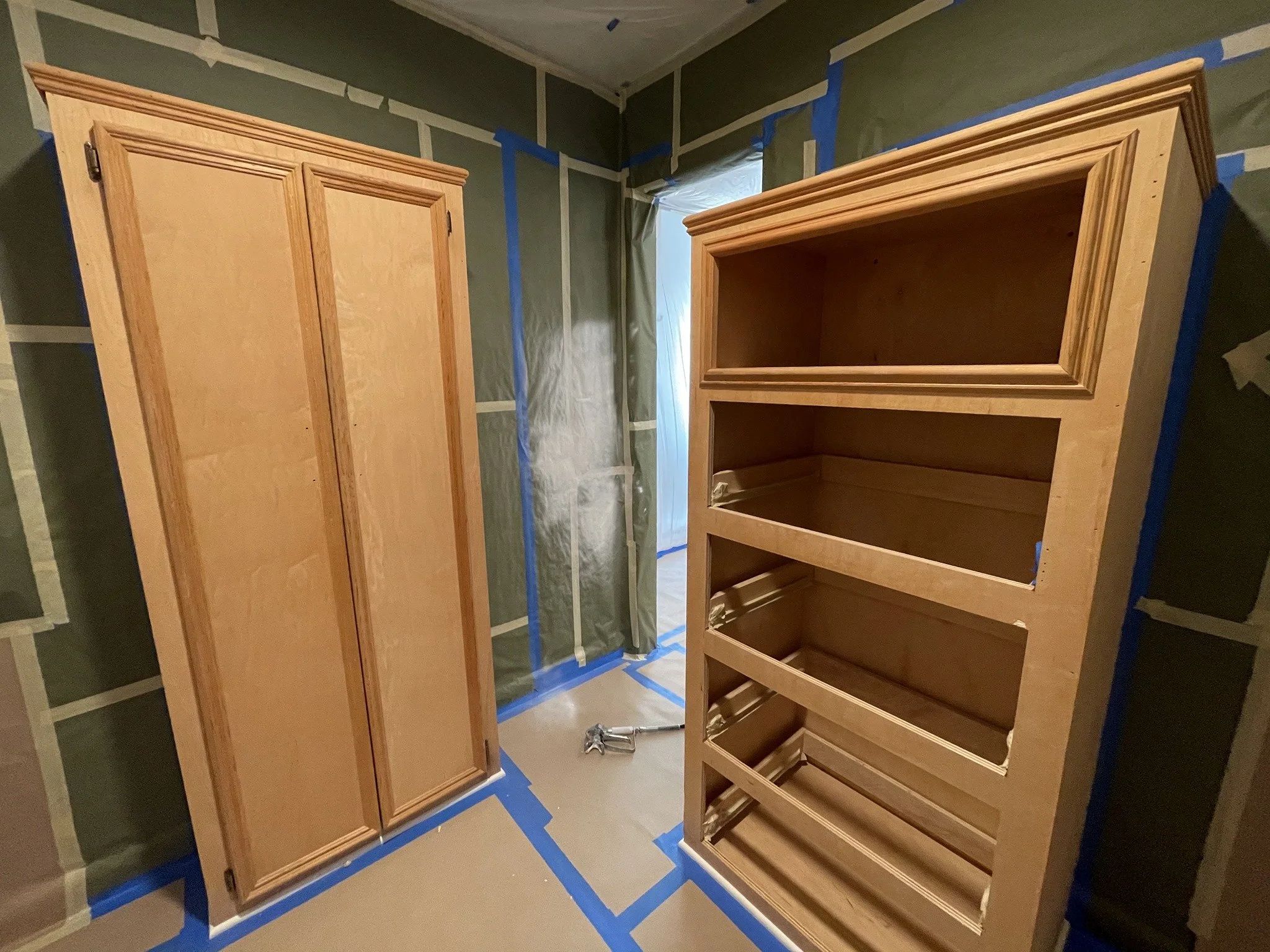 Unfinished wooden cabinet and wooden wardrobe in a room under construction, with drywall and plastic sheeting on the walls, and painter's tape on the floor.