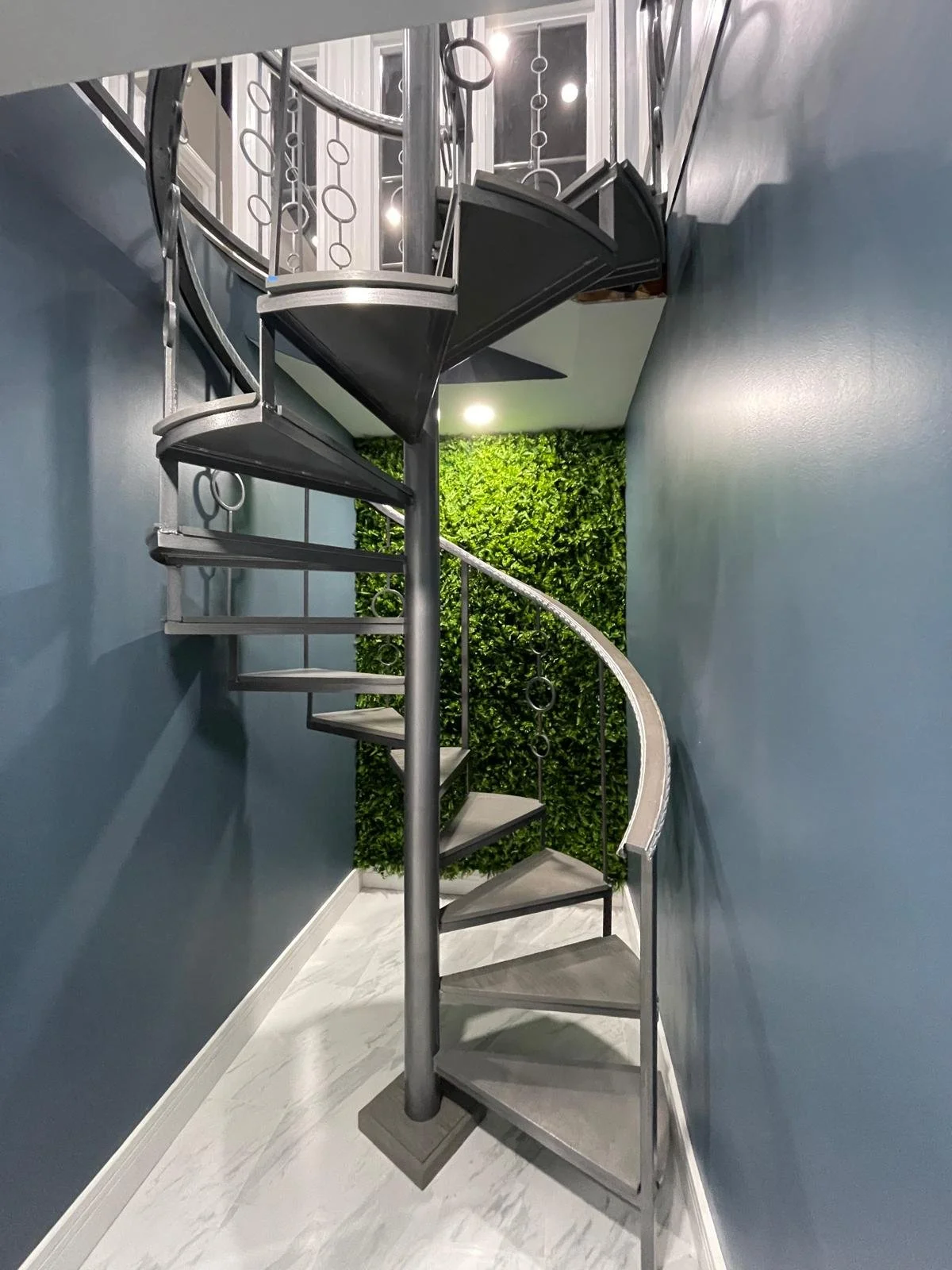 Spiral staircase with metal railings and steps, next to a green wall with plants in an indoor space.