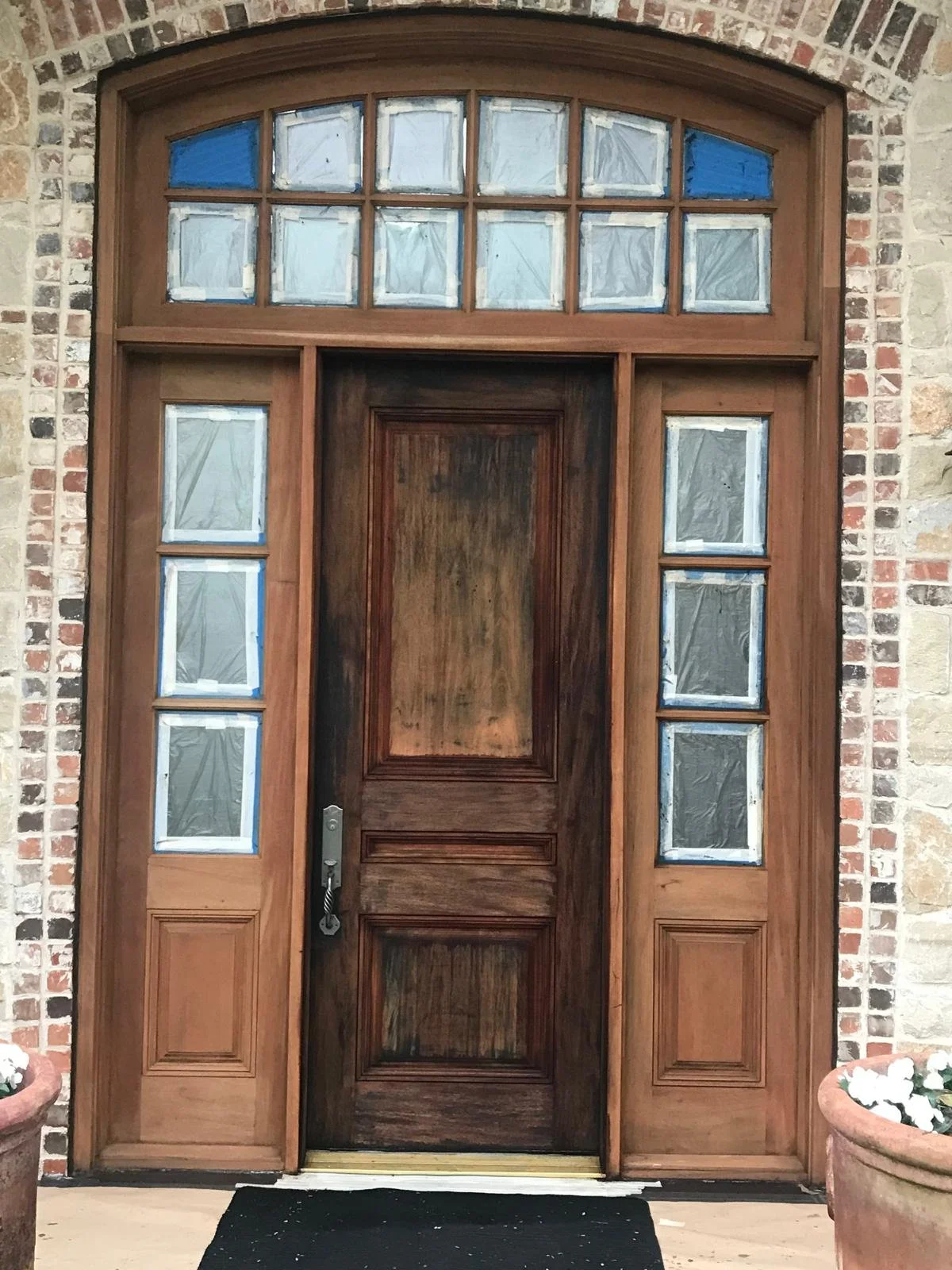 A wooden front door with multiple small window panes, set within a brick wall, with two large planters filled with white flowers on either side.