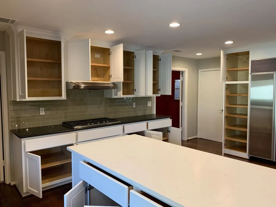 Unfinished kitchen with open cabinets, a black countertop, a stove, a stainless steel refrigerator, a white kitchen island, and a gray backsplash, with furniture and appliances waiting to be installed.