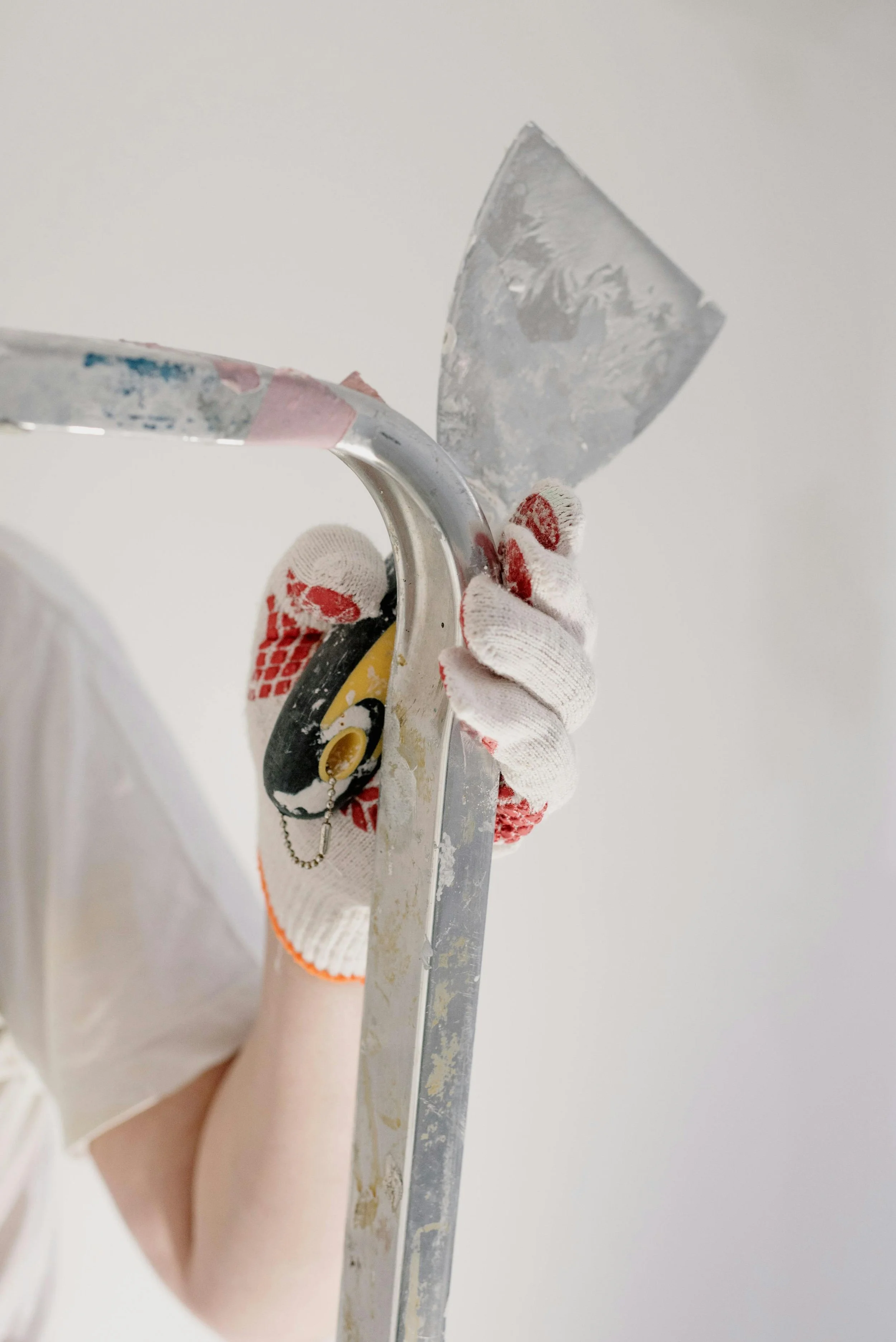 Close-up of a person wearing white gloves while holding a paint scraper and a professional painter's tool, with a plain white wall background.