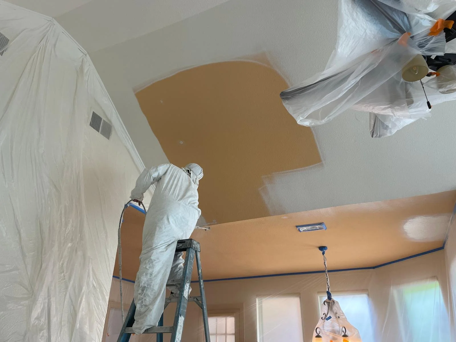 A person wearing protective white coveralls on a ladder painting the ceiling of a room with orange paint, with plastic sheeting covering furniture and walls.
