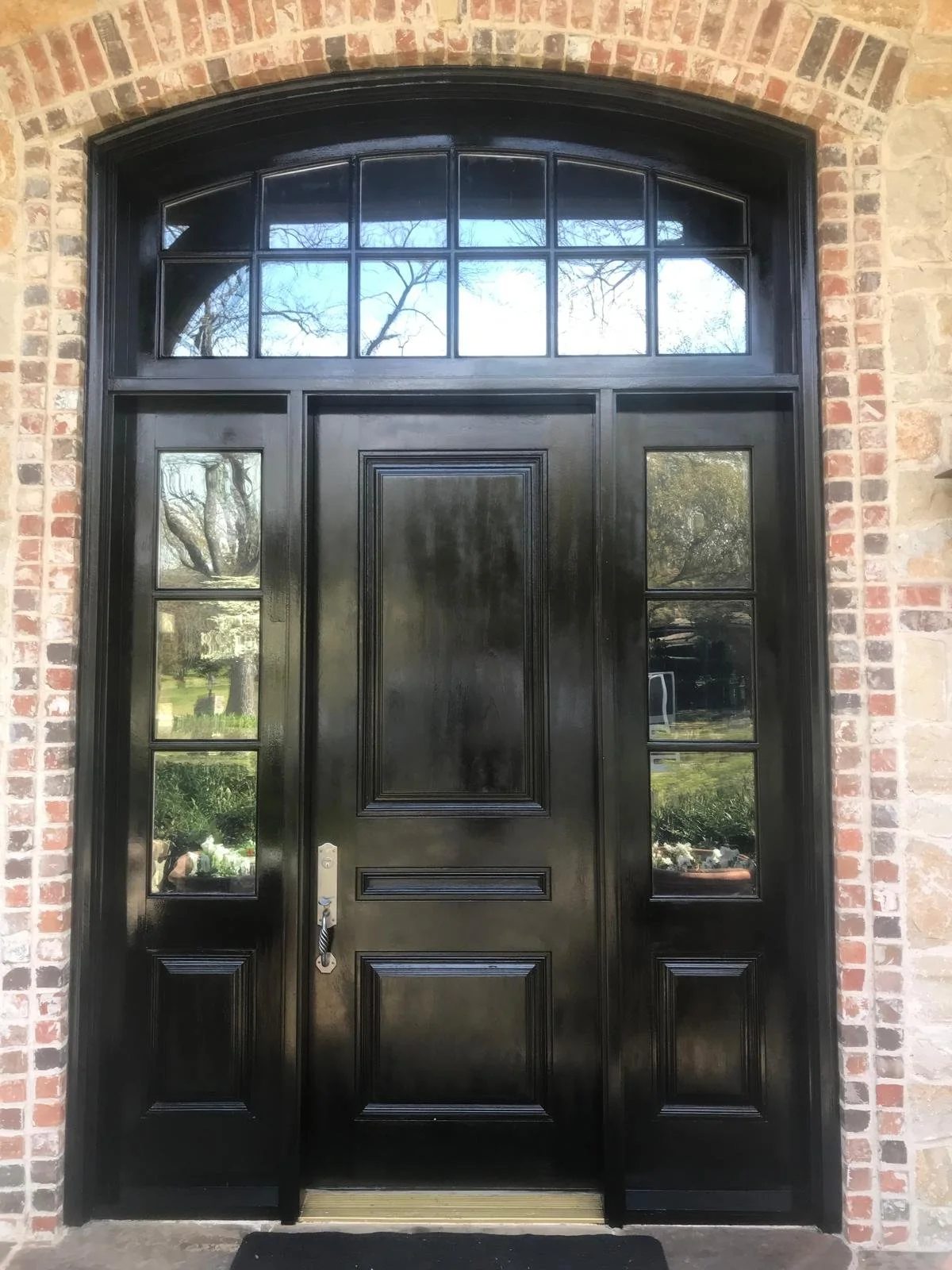 A black front door with decorative panels, flanked by sidelights and topped with a transom window, set into a brick and stone exterior wall.