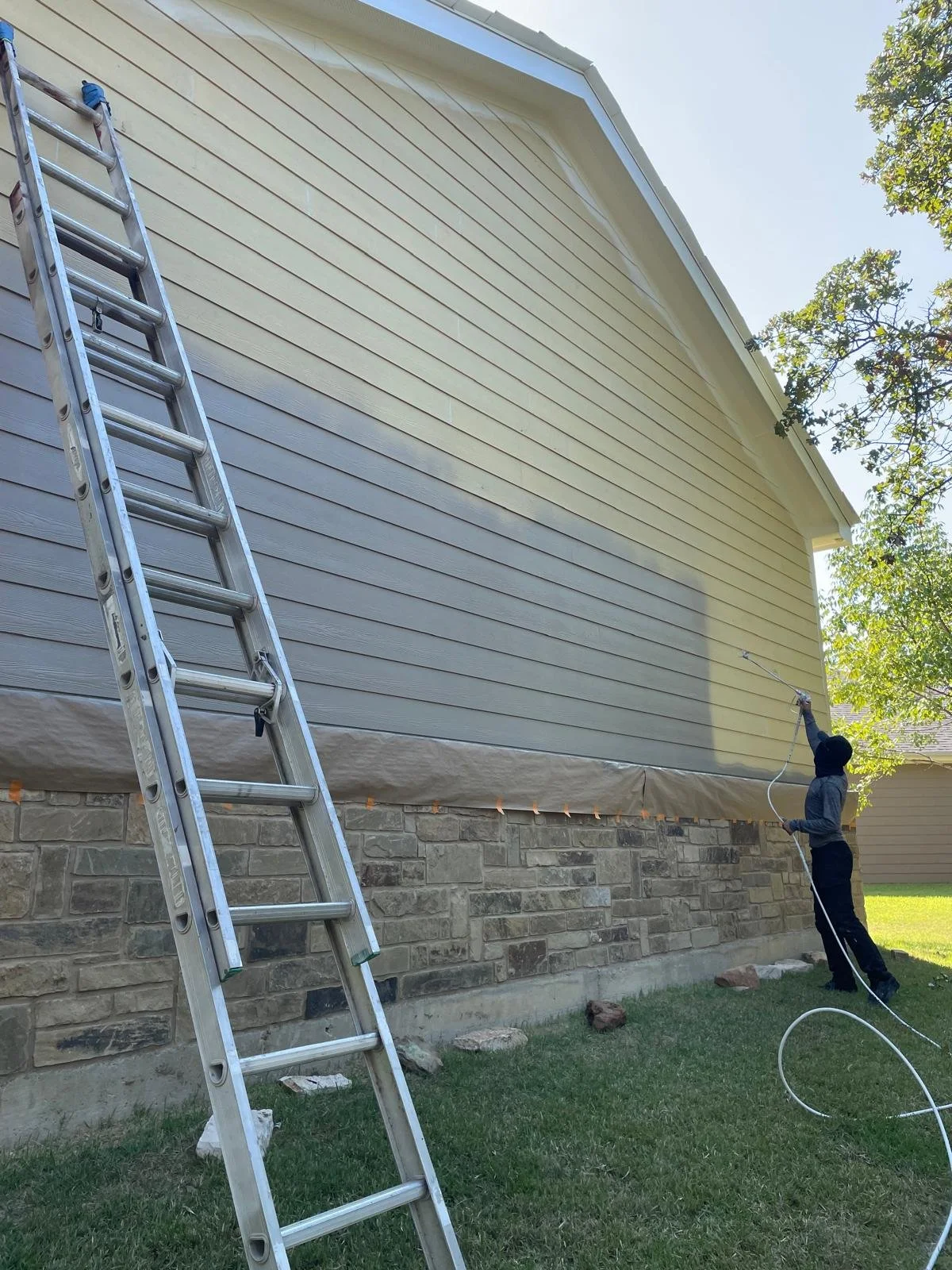 Person power washing the side of a house with beige siding, standing on the ground, using a long wand, next to an aluminum ladder leaning against the house near a stone foundation, in a yard with grass and a tree with green leaves.