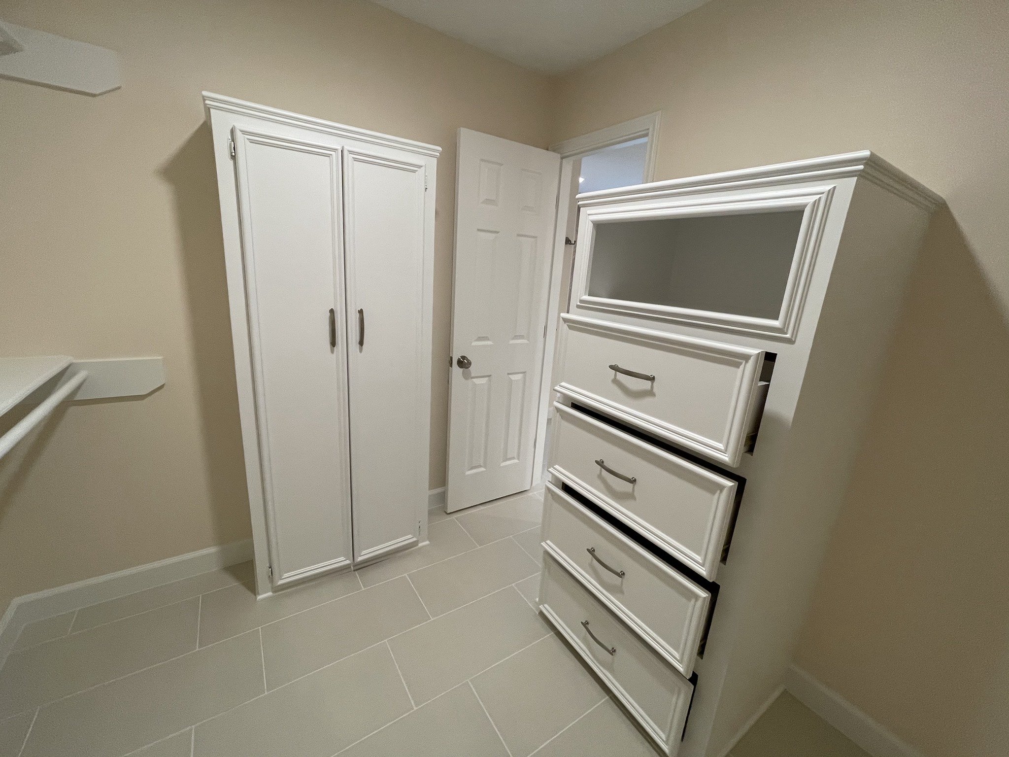 A small room with beige walls and light-colored tiled floor. Contains a white wardrobe, a white dresser with multiple drawers, and a white door slightly open. There is also a small white shelf on the wall to the left.