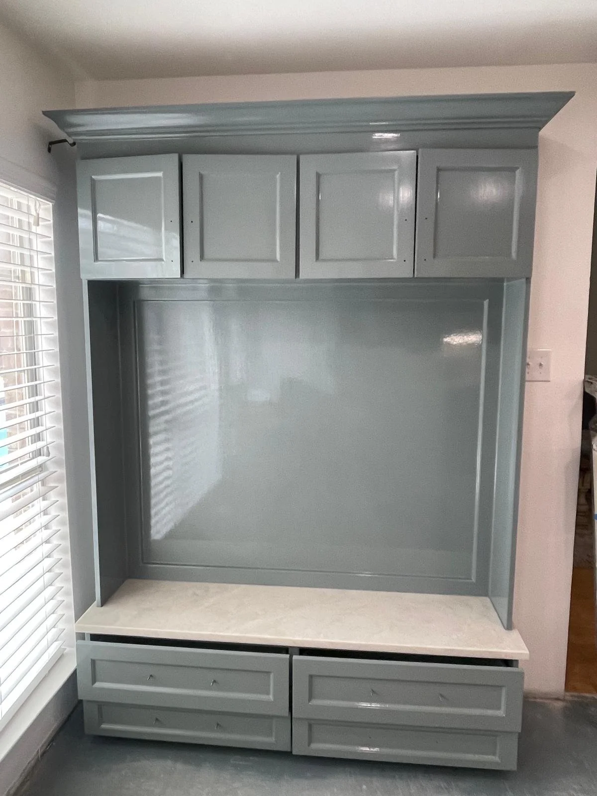 A painted grey built-in entertainment center with upper and lower cabinets, a central open space, and a white countertop at the bottom, located beside a window with blinds.