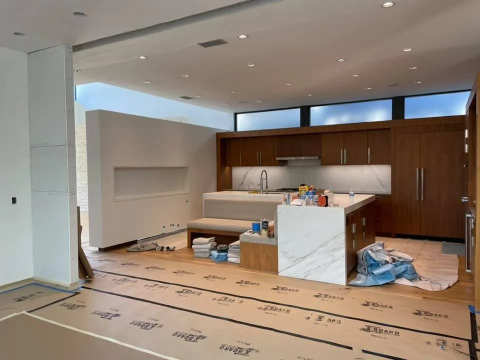 Modern kitchen under construction with wooden cabinets, white marble countertops, and a kitchen island. Construction materials and tools are on the island and floor.